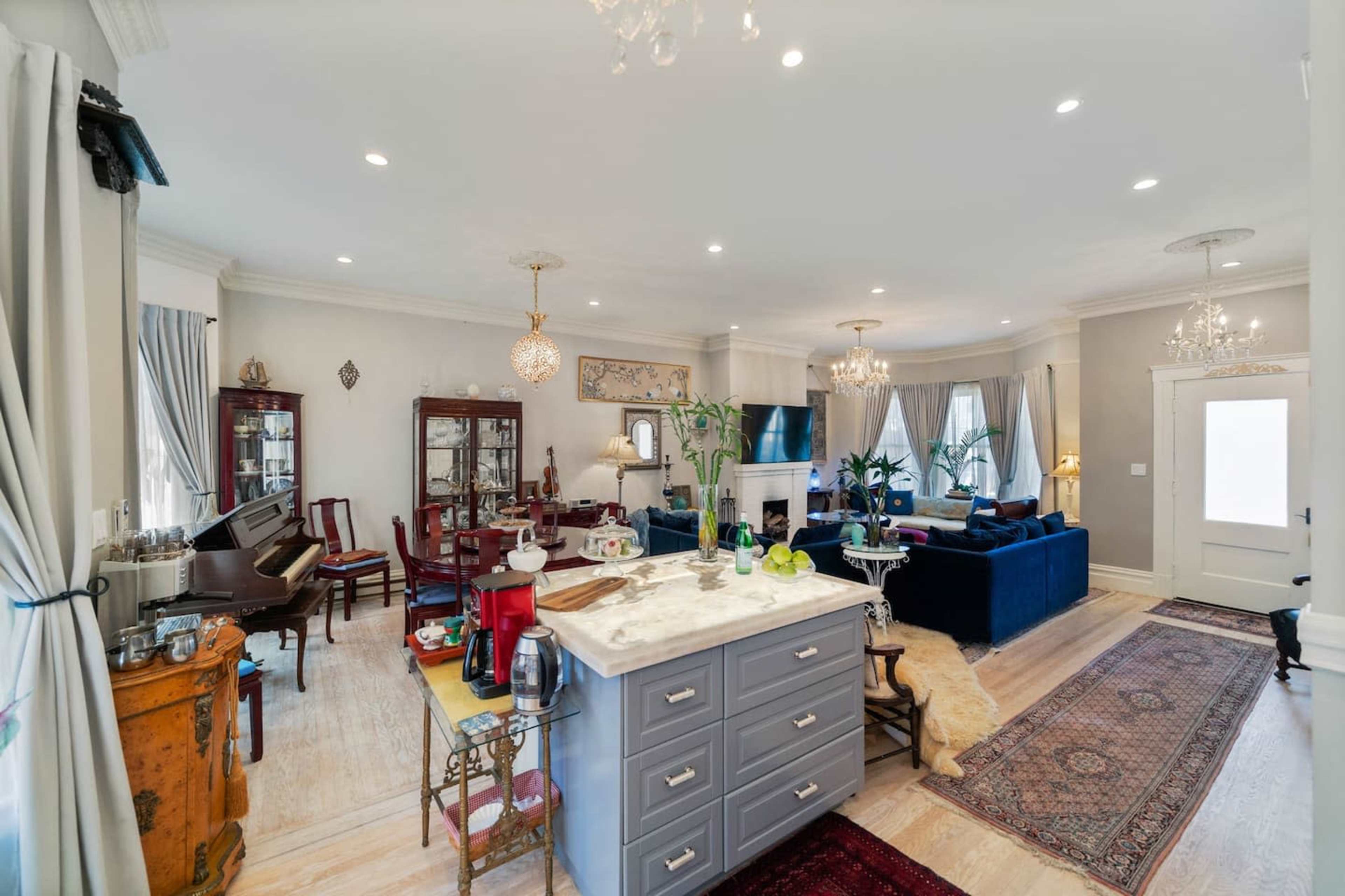 A spacious living area featuring a kitchen island, a seating area with a dark sofa, and a piano in one corner, all complemented by elegant lighting and decor.