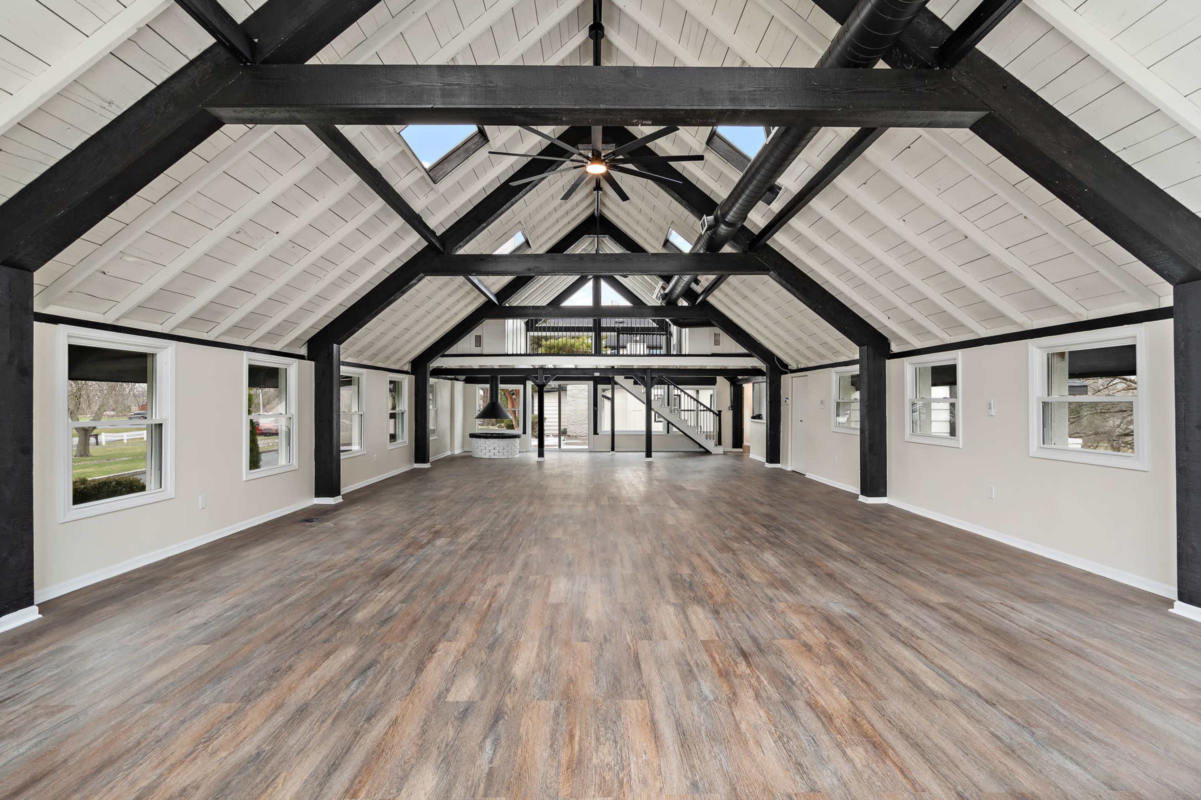 The image shows a spacious, open interior room with a high, beamed ceiling and large windows, featuring wooden flooring.