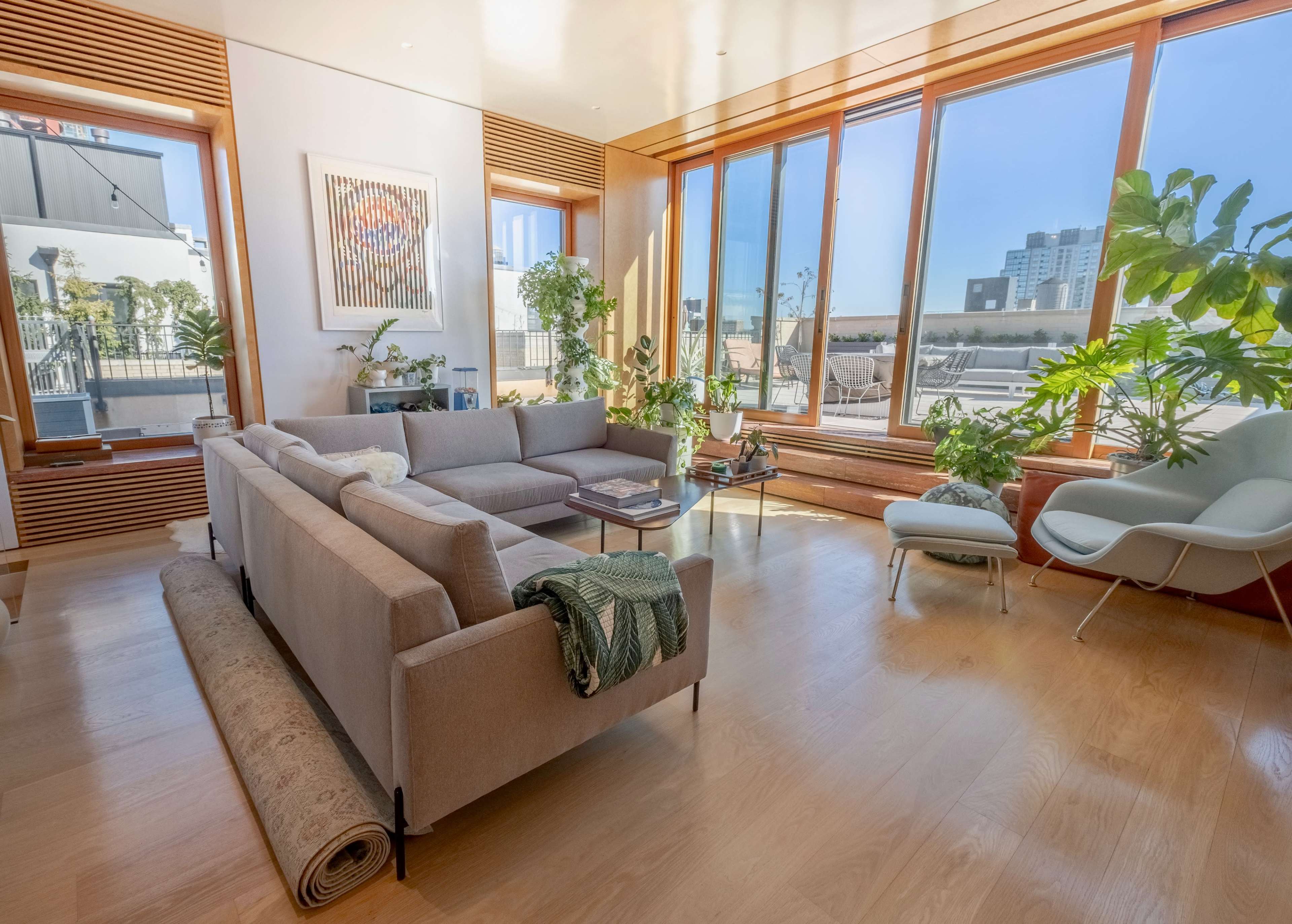 A bright living room features a large sofa, potted plants, and expansive windows with a view of the city.