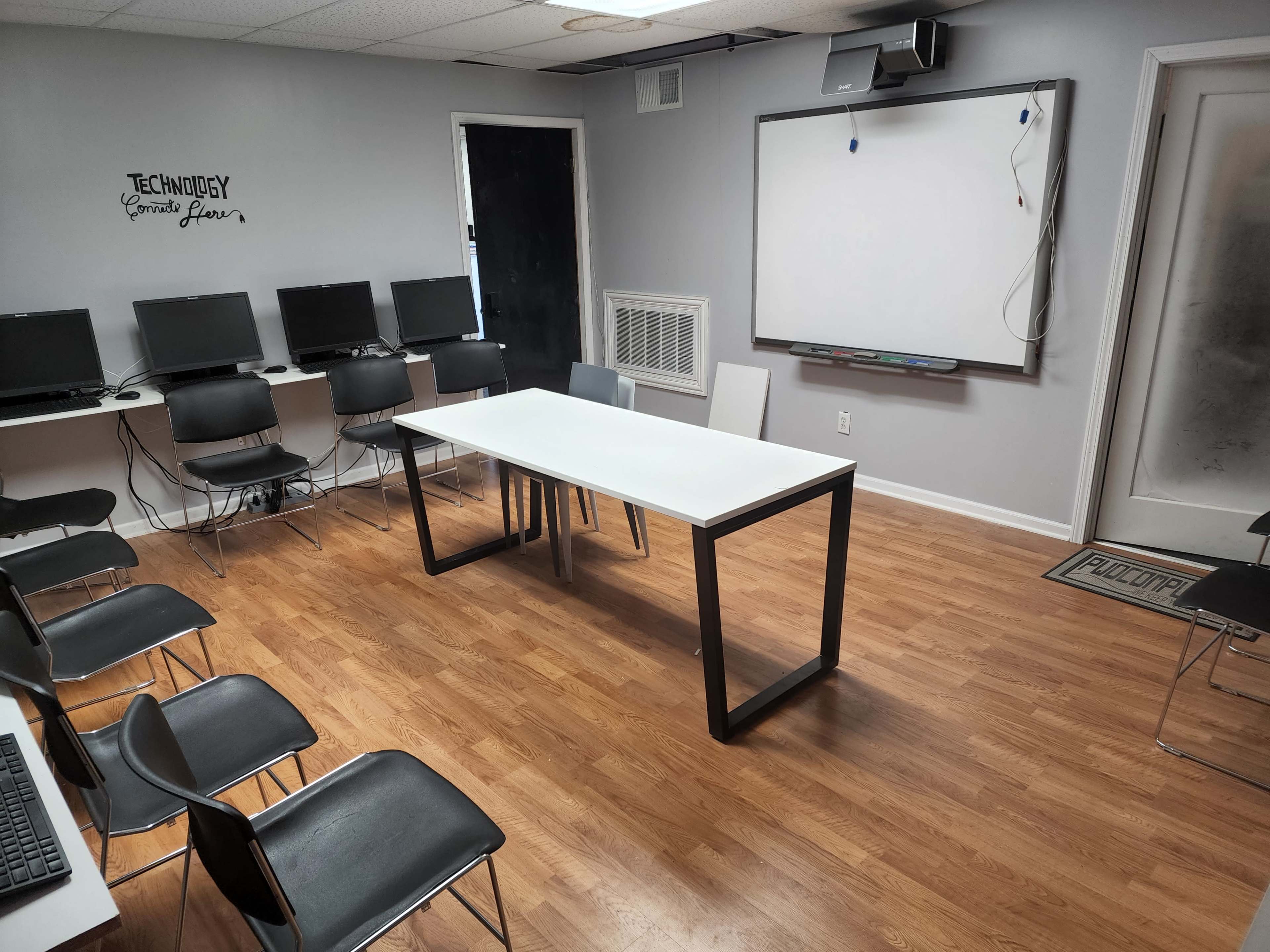 A classroom features a rectangular table at the center, surrounded by black chairs, with computers and a whiteboard on the walls.
