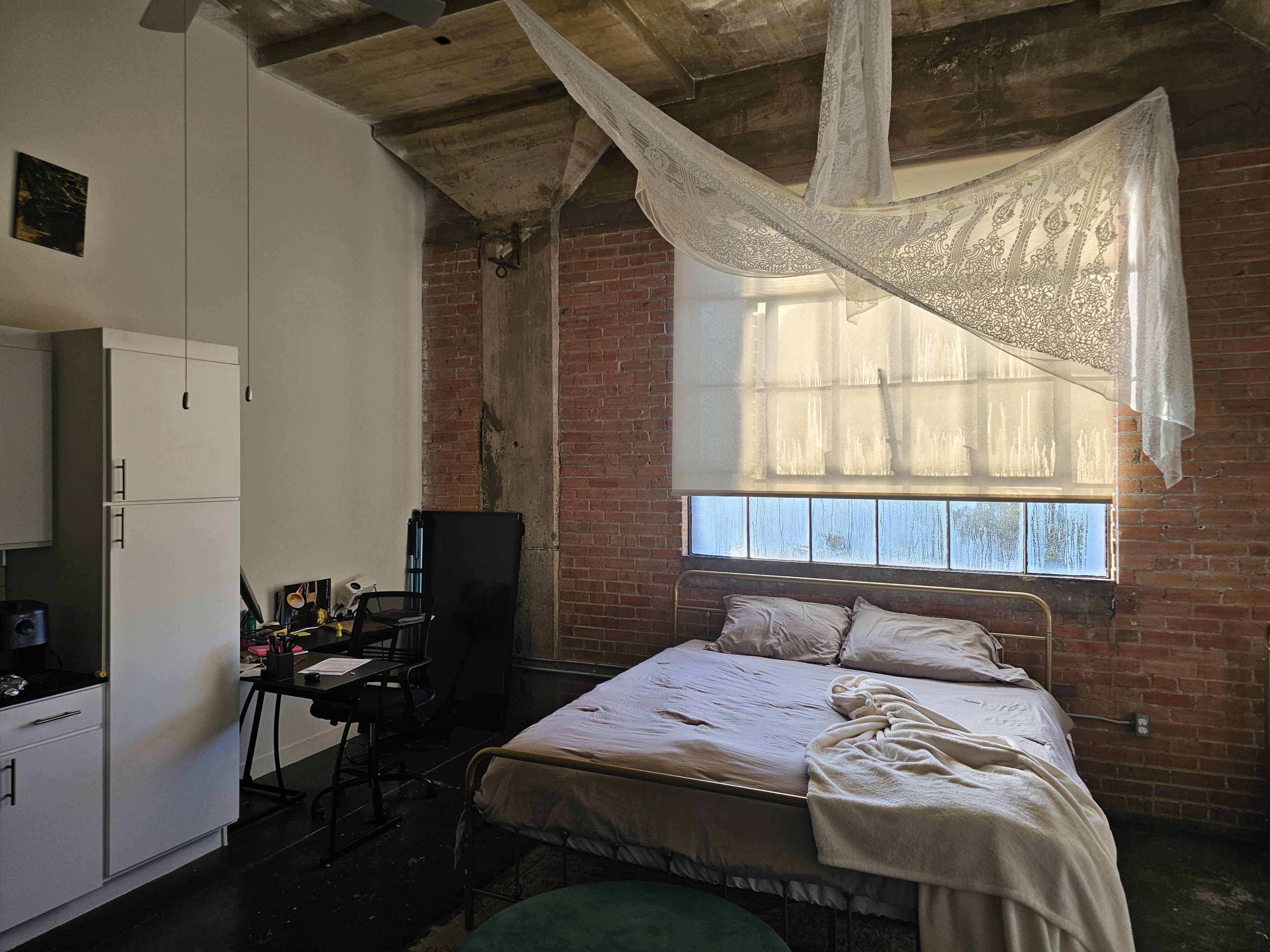 The image shows a bedroom with a bed, a desk with a chair, a refrigerator, and a large window covered with a sheer curtain, set against exposed brick walls and a concrete ceiling.