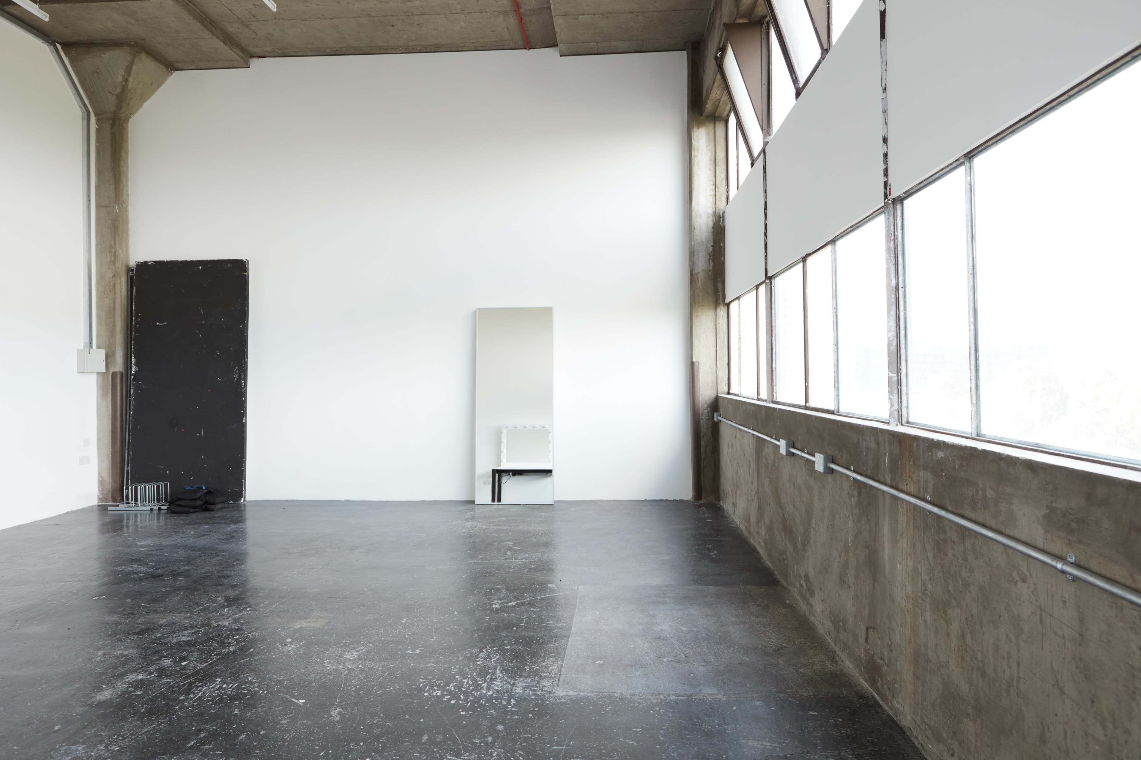 A spacious, empty room with large windows, a concrete wall, and a mirror positioned on one side.