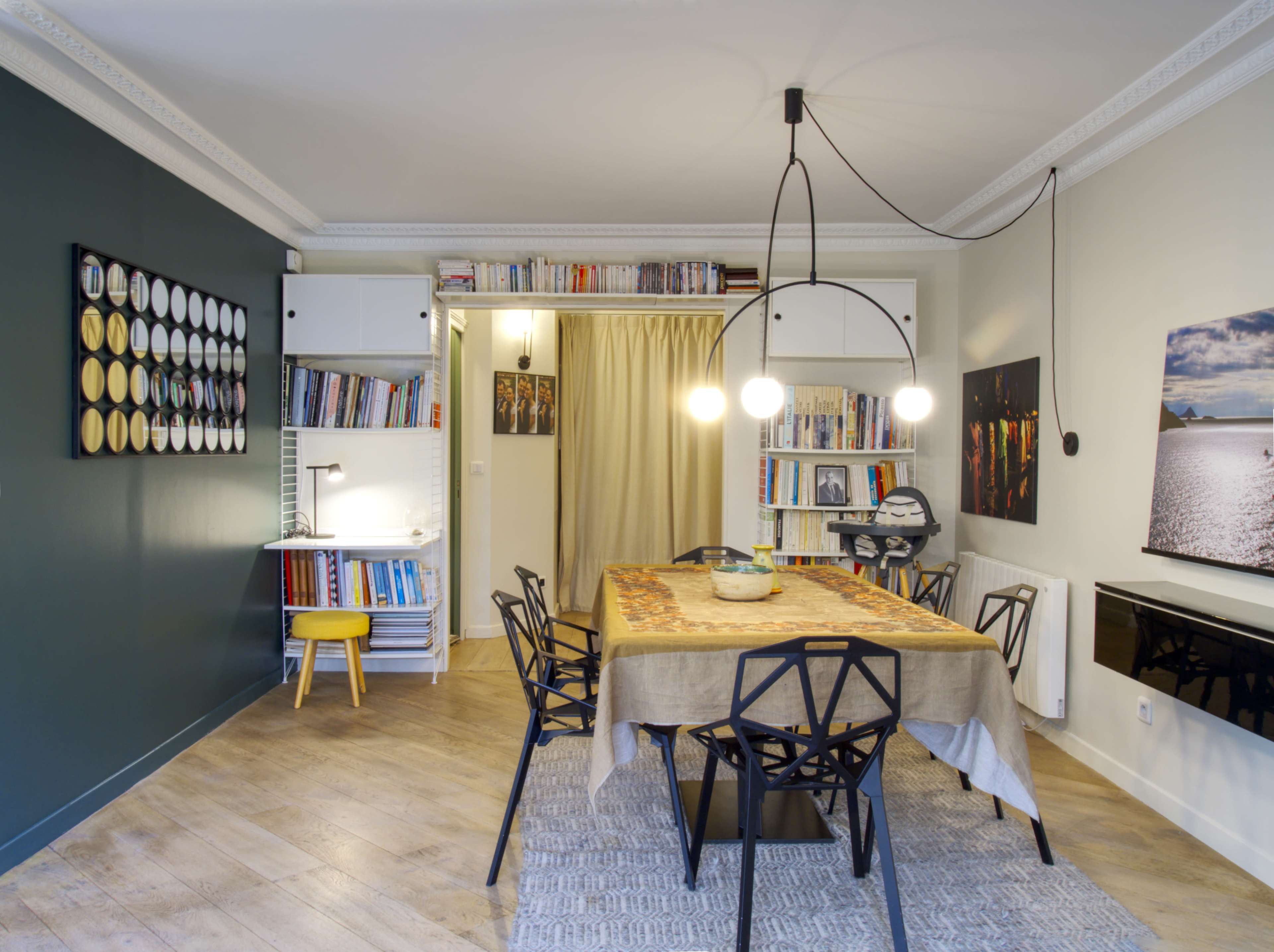 The image shows a modern dining area featuring a central table with black chairs, a bookshelf, and decorative wall art.
