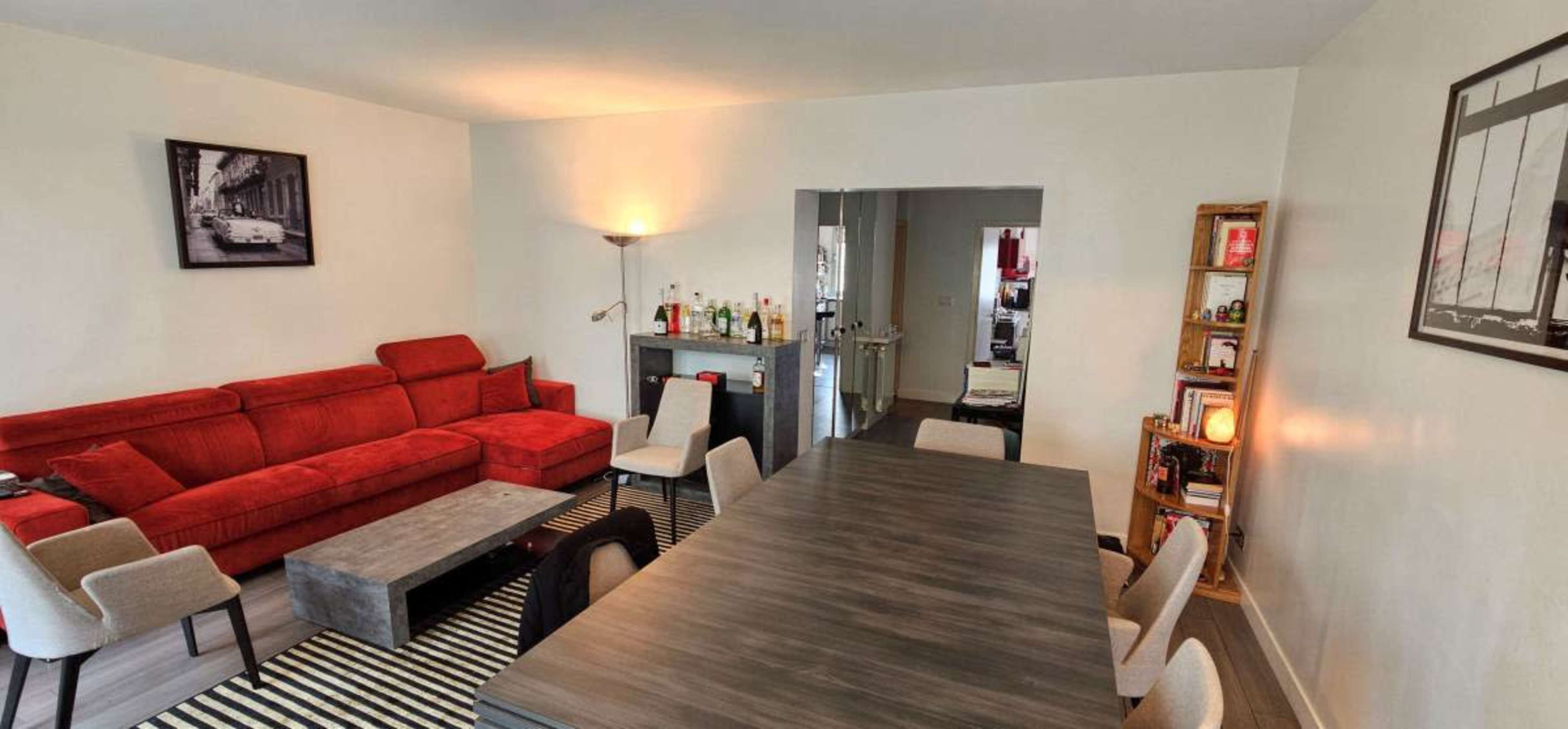 The image shows a spacious living and dining area with a red sectional sofa, a wooden dining table surrounded by chairs, and a bar area with bottles on the side.