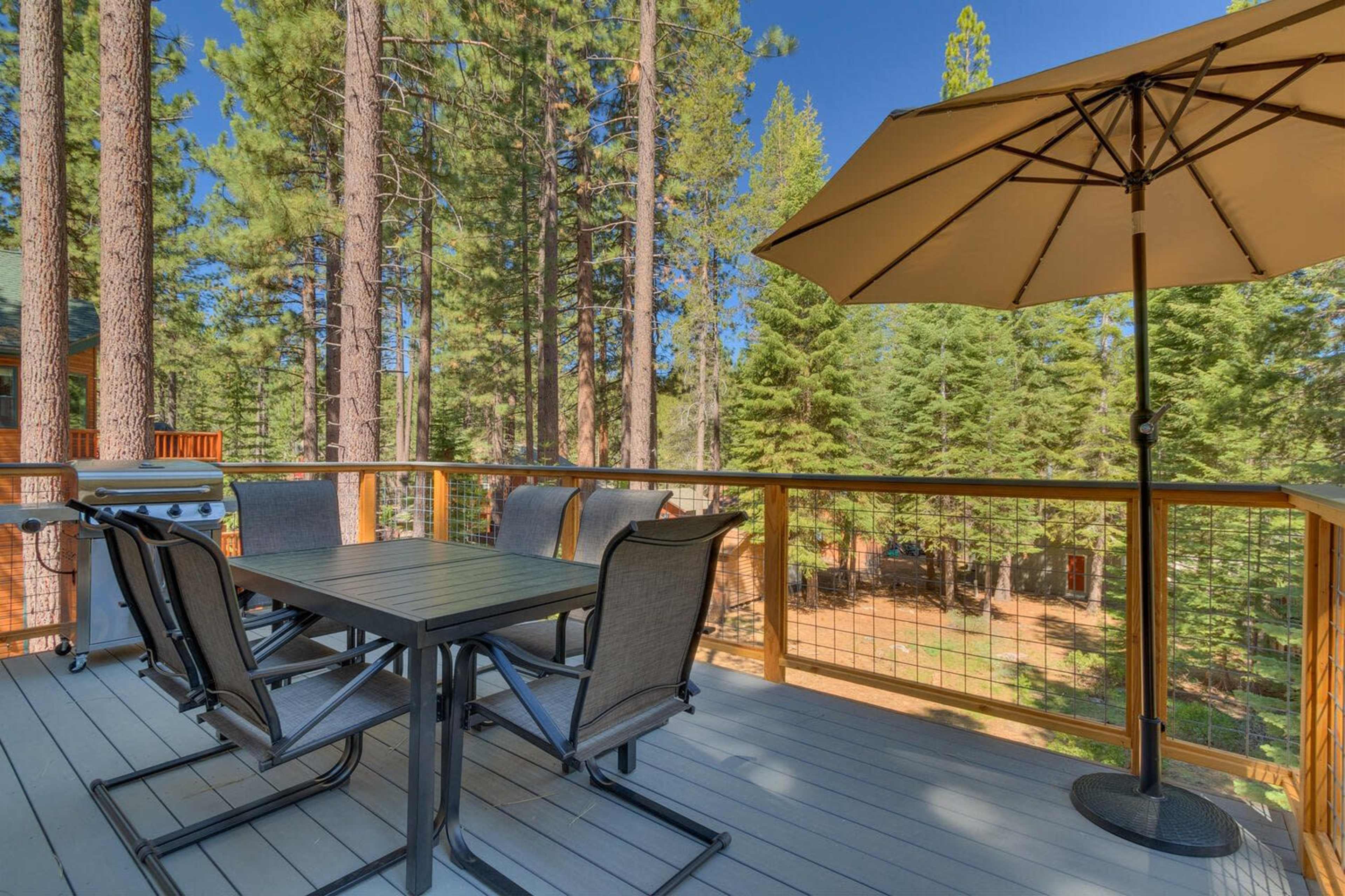 A wooden deck features a dining table with six chairs and an umbrella, surrounded by tall trees in a forested area.