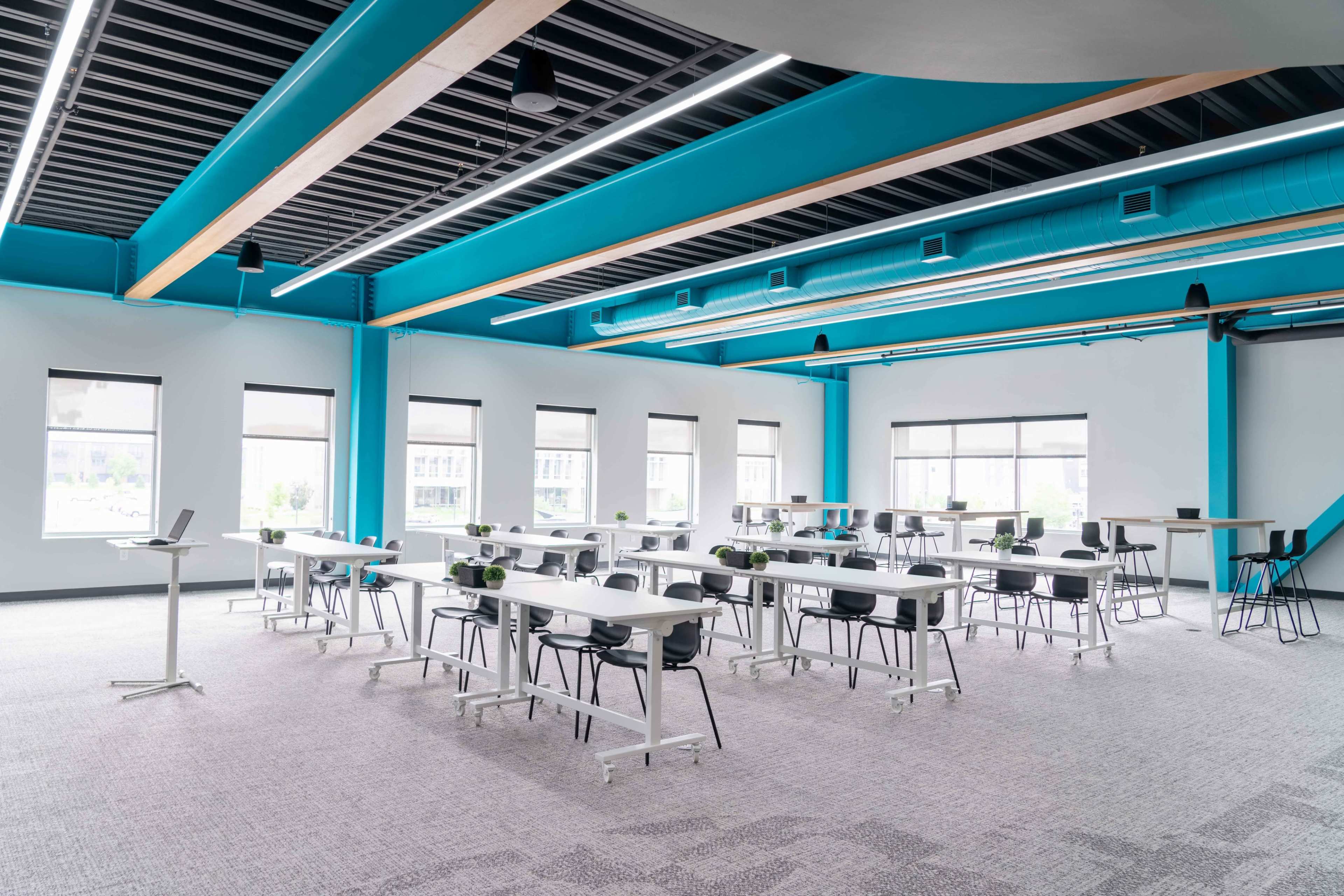 The image shows a modern, brightly lit training room with several long tables arranged in a layout for meetings, surrounded by black chairs and green plants on the tables.