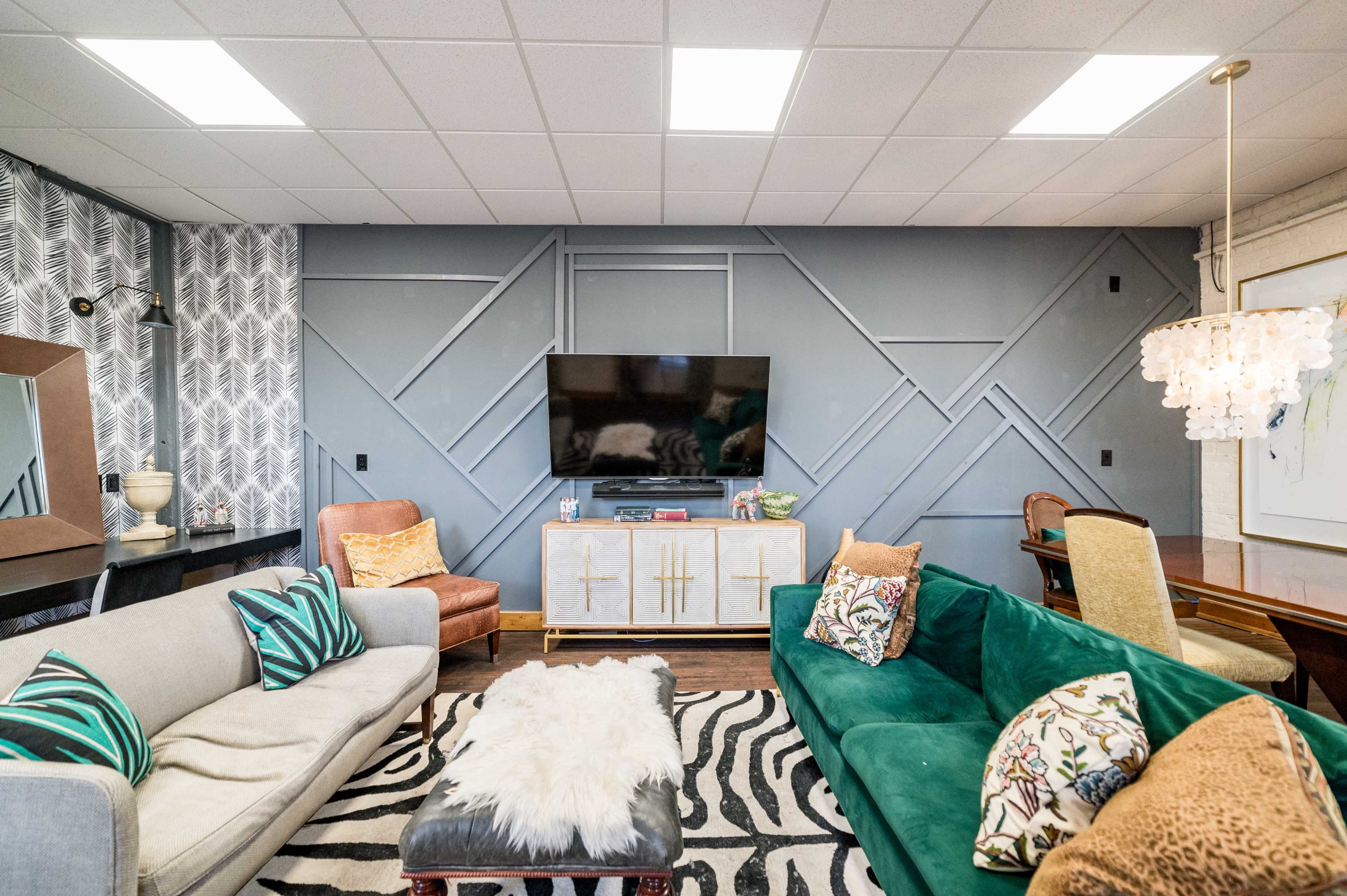 The living room features a sofa, a green velvet couch, a television mounted on a geometric accent wall, and a large area rug with a zebra pattern.