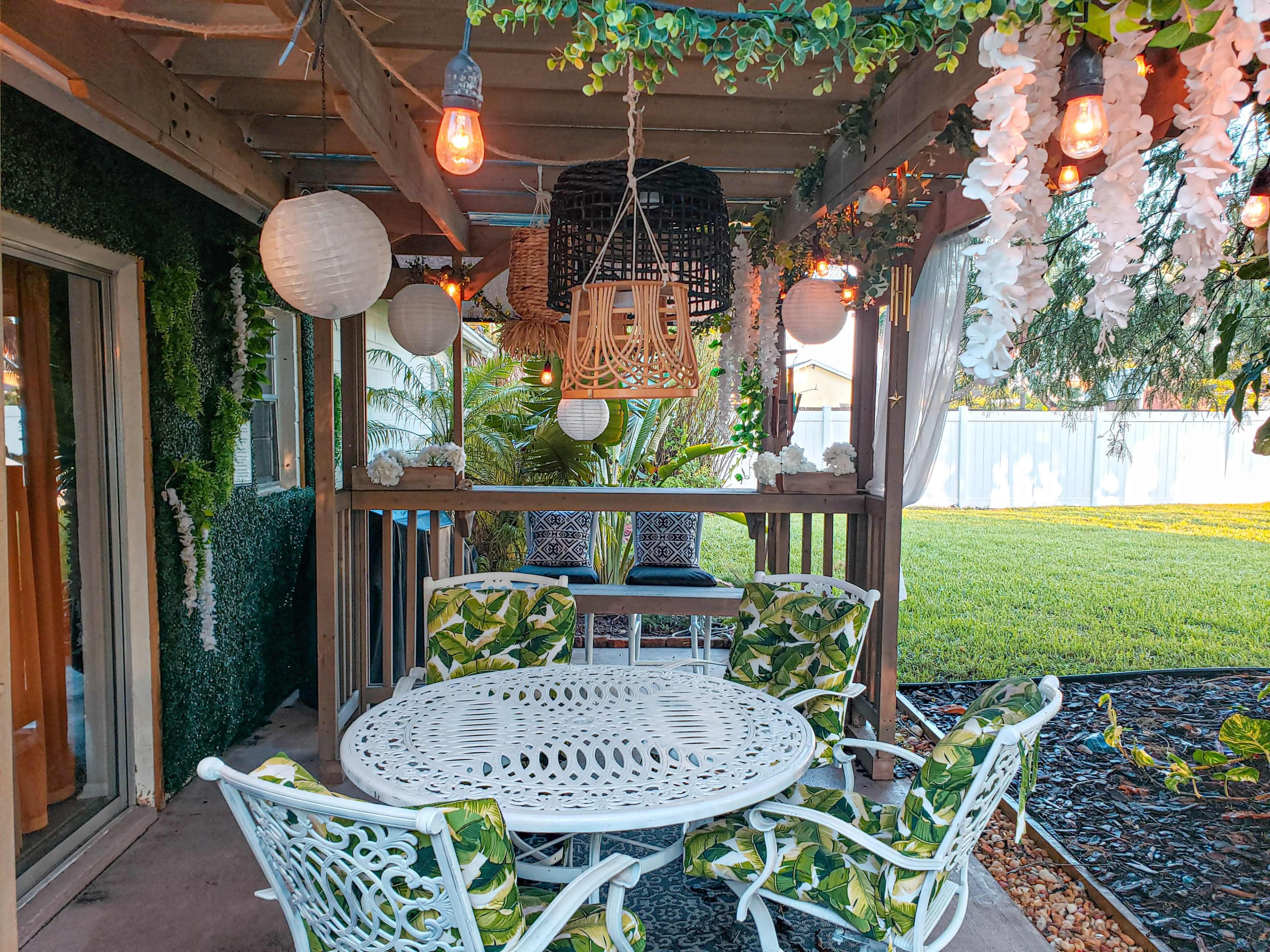 A patio area with a round table and four chairs, surrounded by greenery and decorated with hanging lanterns and floral accents.