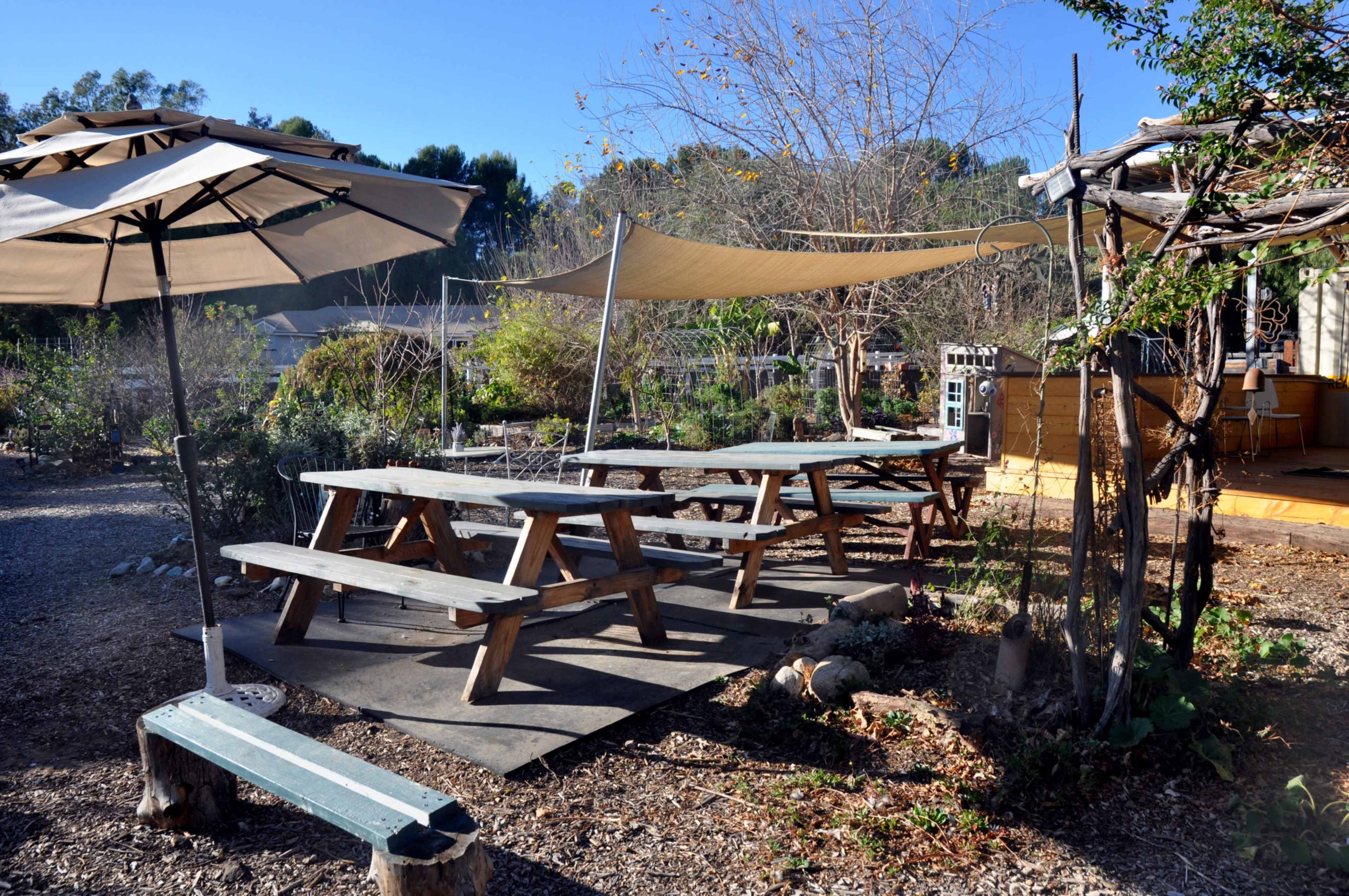 The image shows several wooden picnic tables under shaded umbrellas in a garden setting.