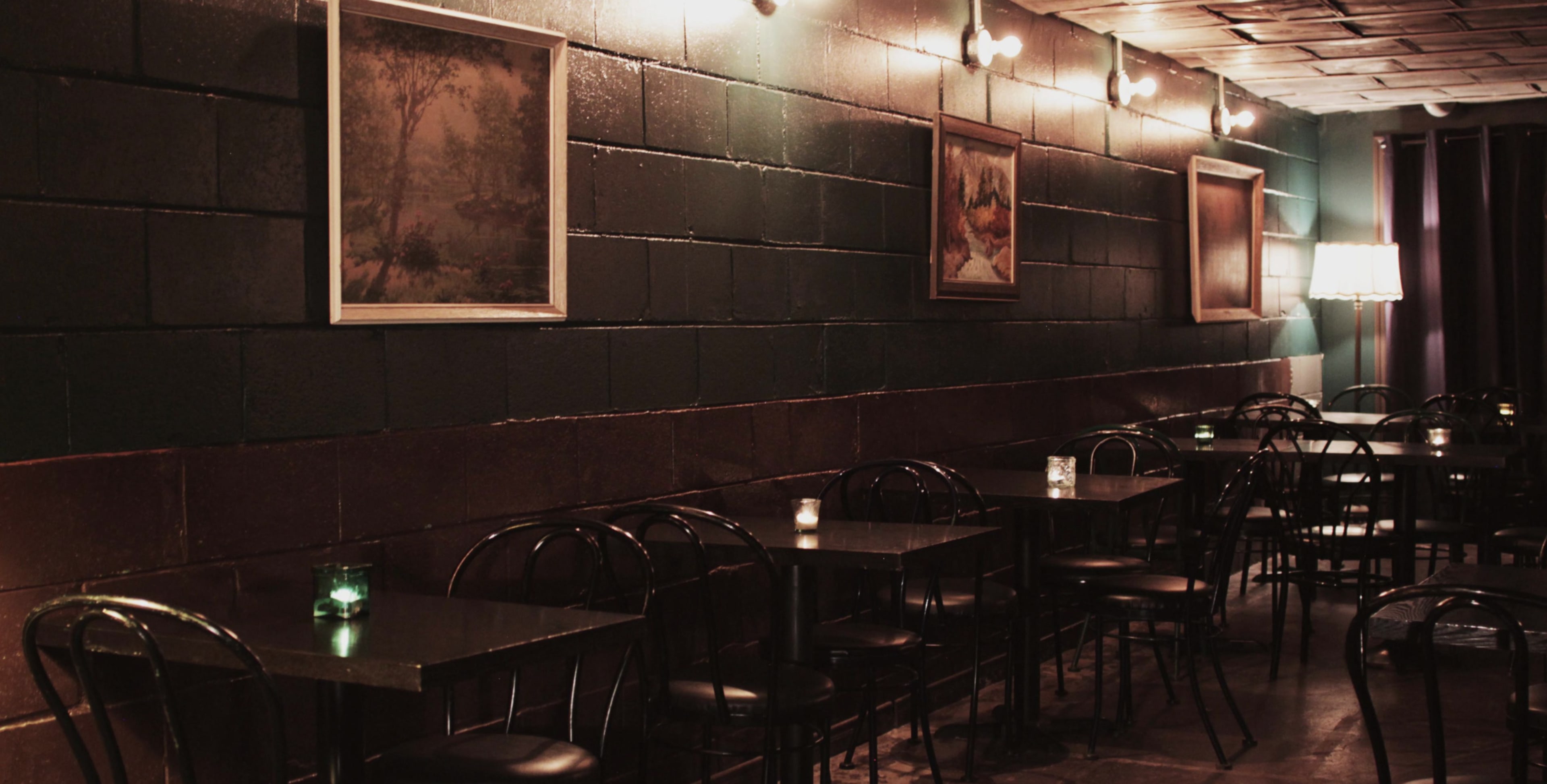 A dimly lit café interior with black tables and chairs, framed paintings on the walls, and small candles on the tables.
