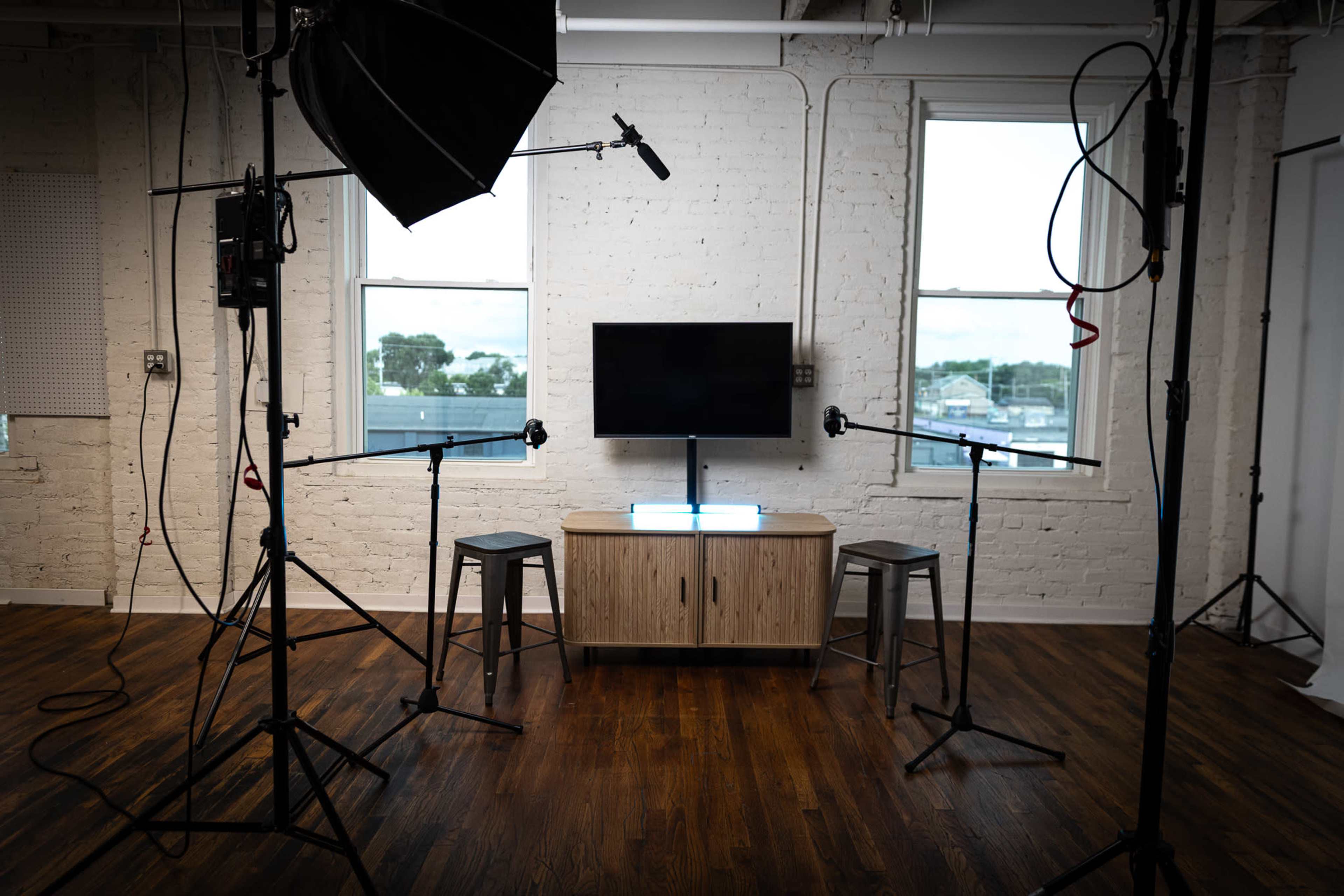 The image shows a minimalistic studio setup with two tall stools, a television on a wooden cabinet, and several lighting and microphone equipment arranged around the space.