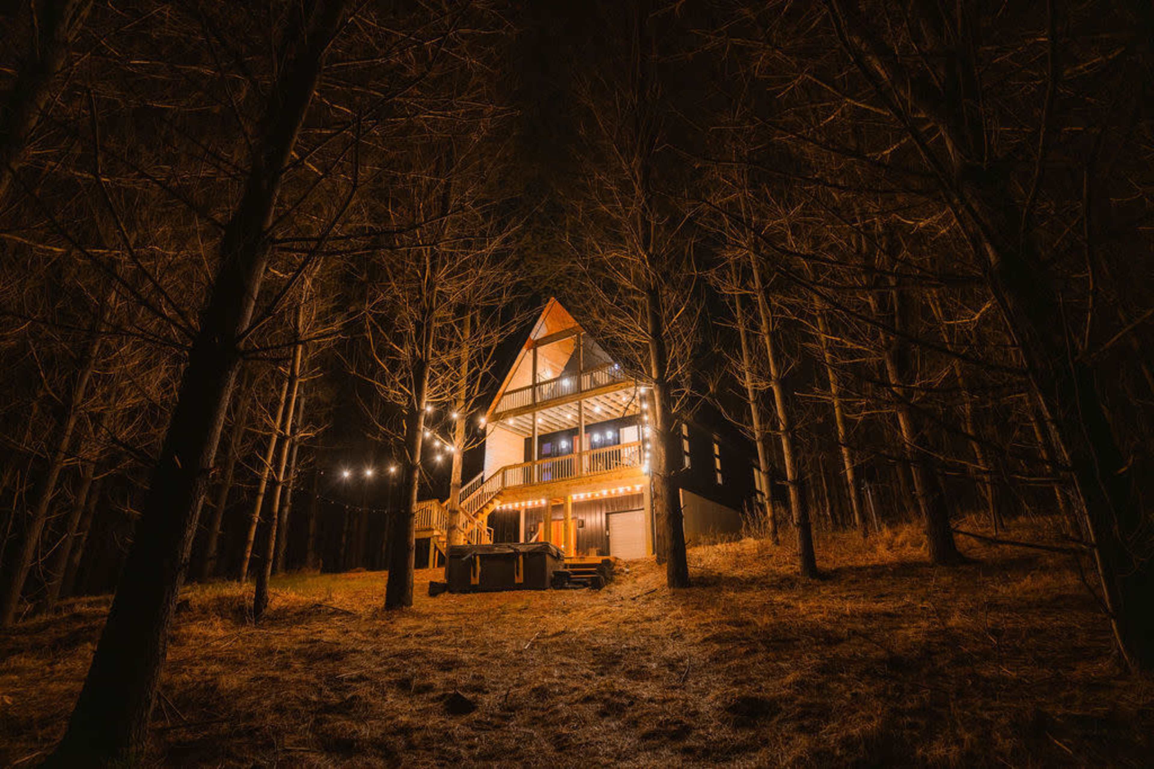 A wooden A-frame house is illuminated by string lights and stands among tall, bare trees at night.
