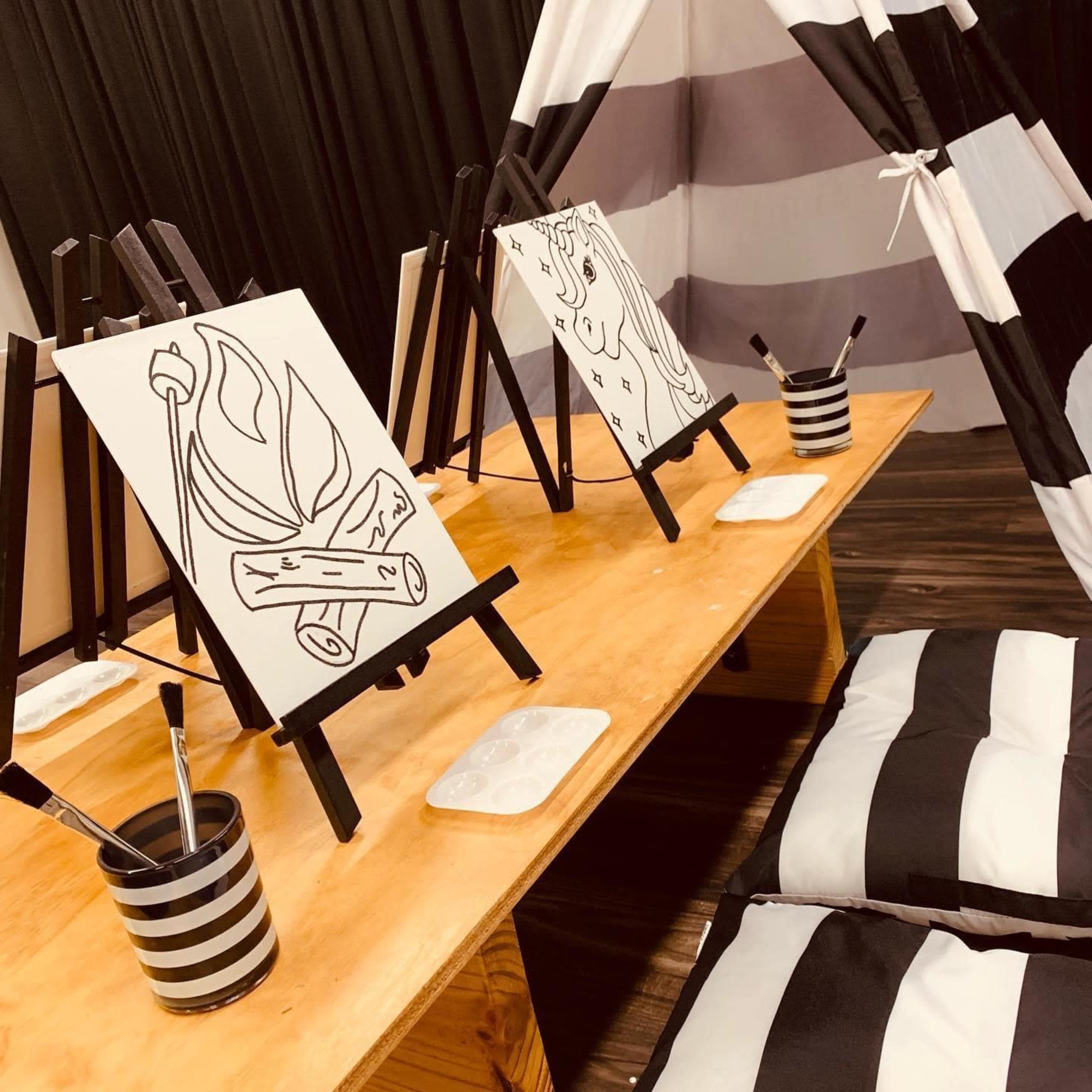 The image shows a table with two blank canvases on easels, paintbrushes, and a palette, set up inside a striped tent.