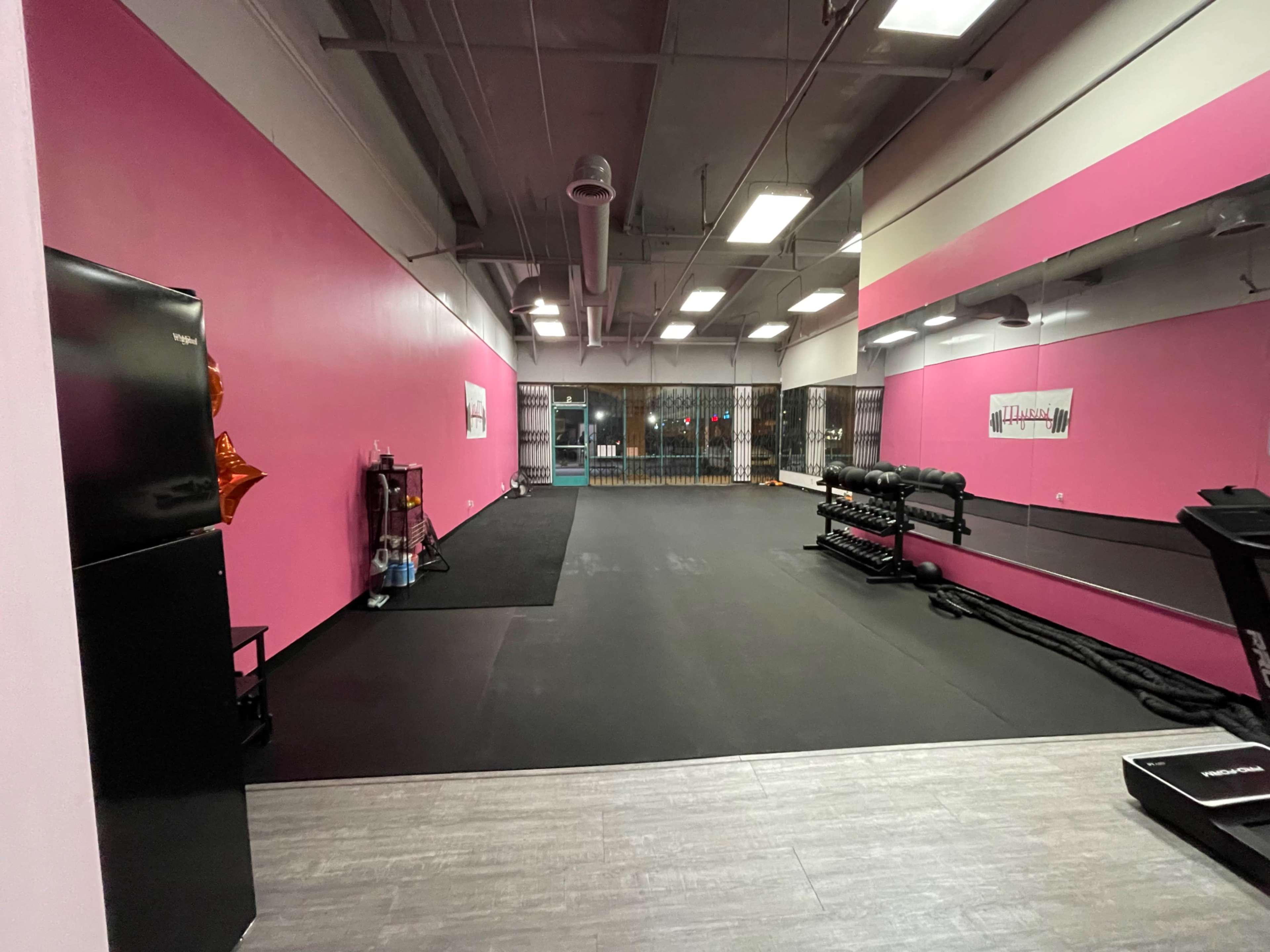 The image shows a fitness studio with pink walls, black flooring, and various exercise equipment positioned along one side.