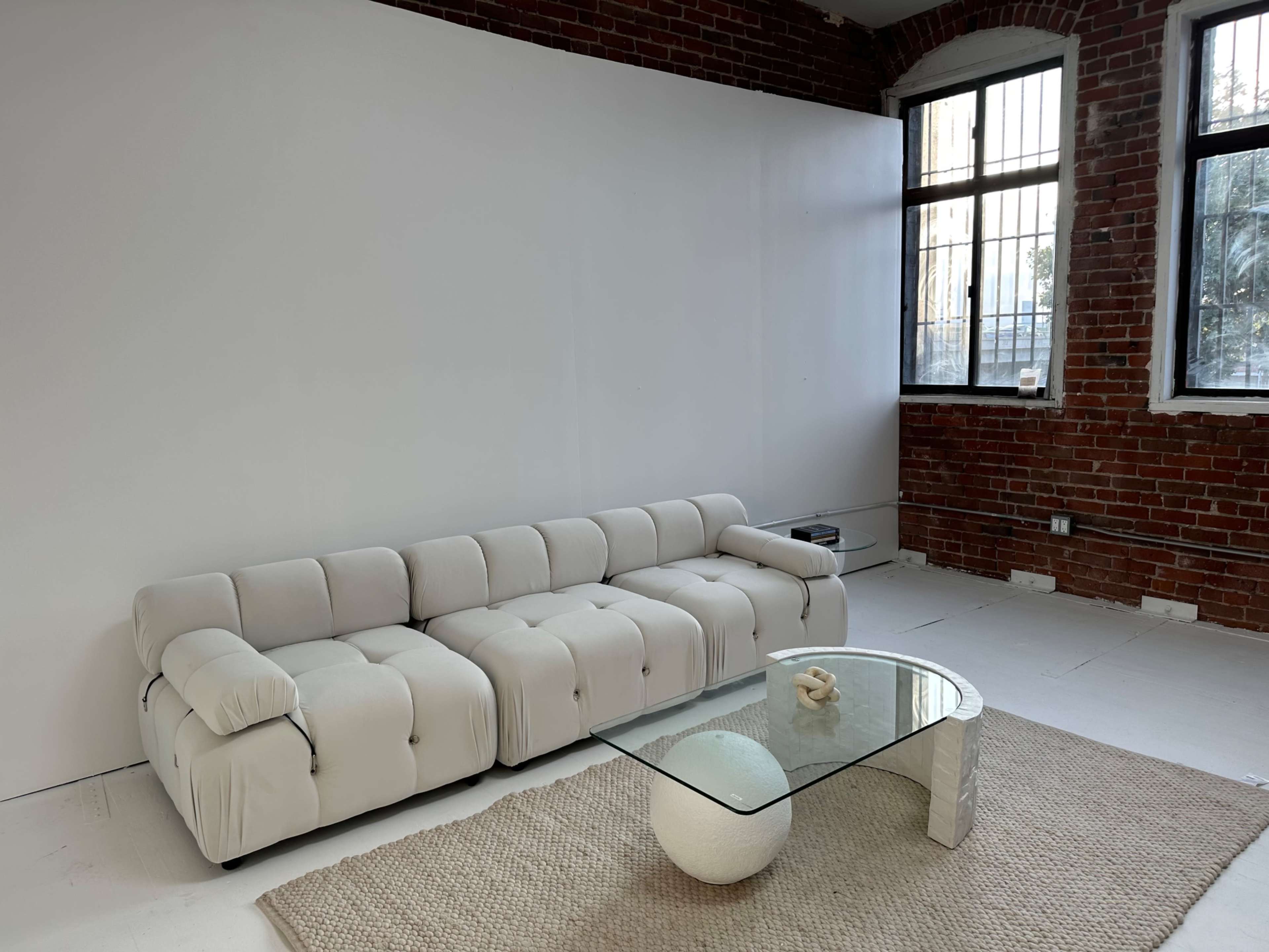 A modern white sofa with a minimalist glass coffee table on a textured rug in a room with exposed brick walls and large windows.