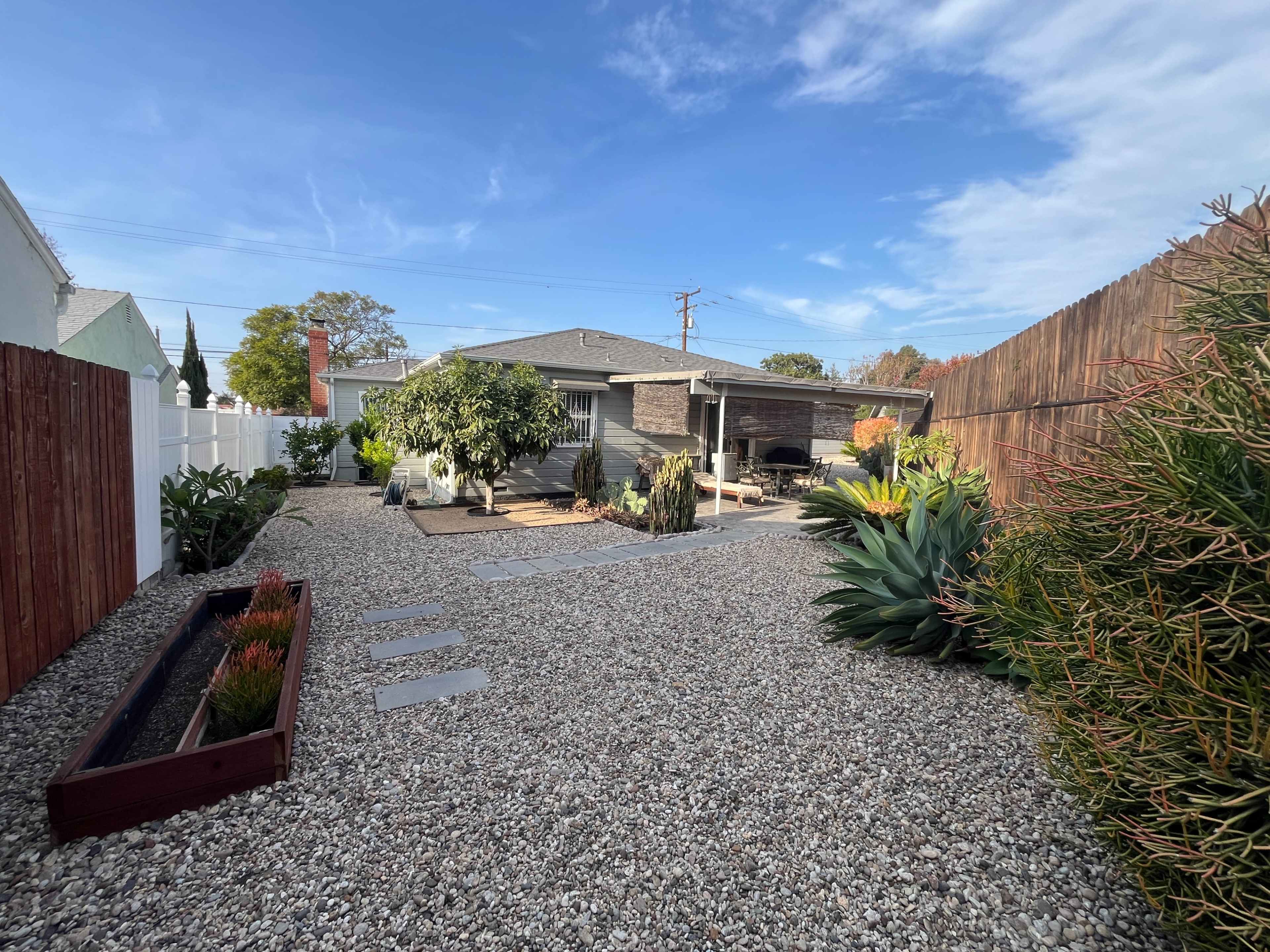The backyard features a gravel landscape with potted plants, a covered seating area, and a shed in the background.