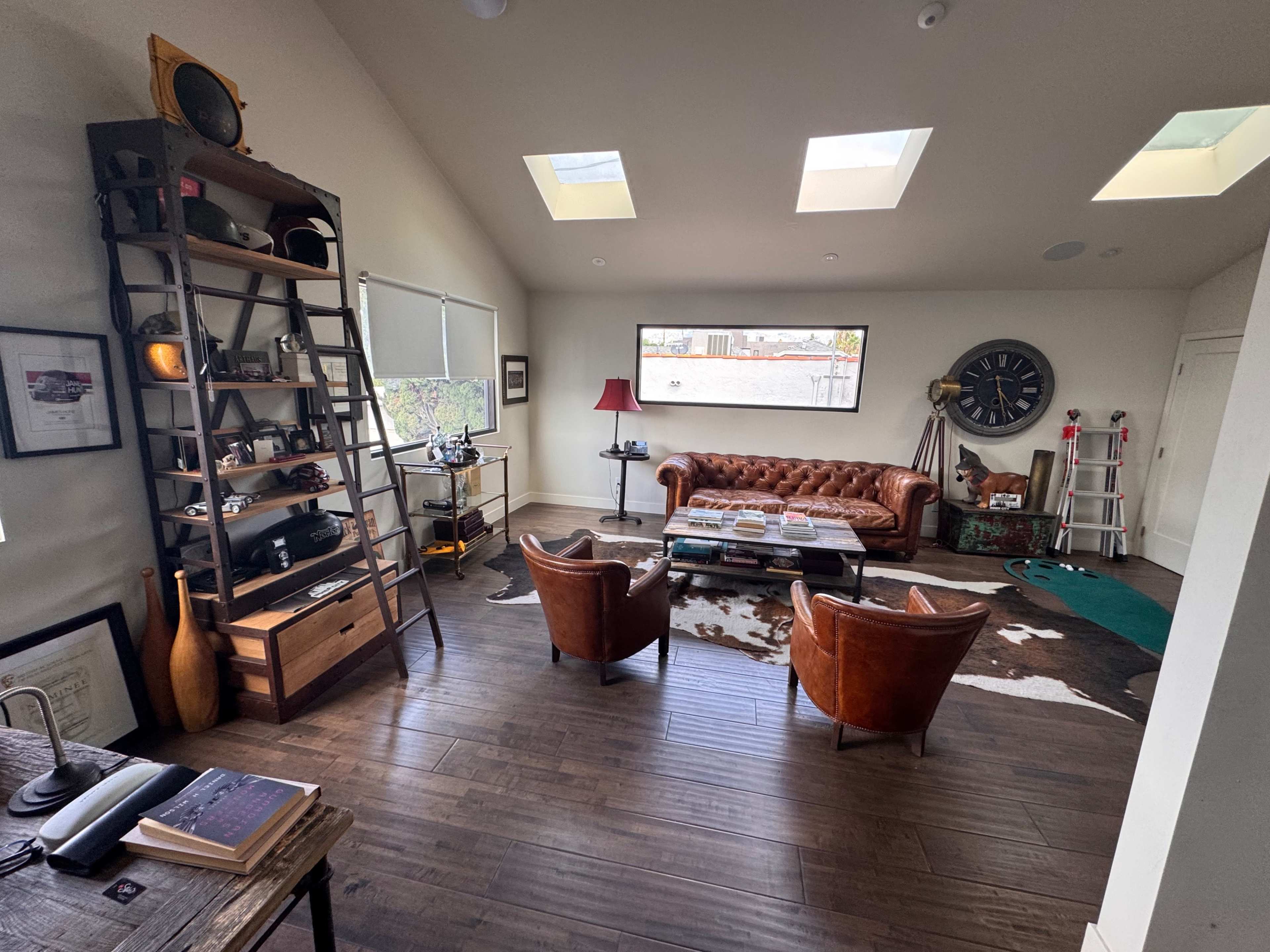 The image shows a spacious, well-lit room featuring a leather couch and two armchairs, a wooden bookshelf, and large windows with skylights.