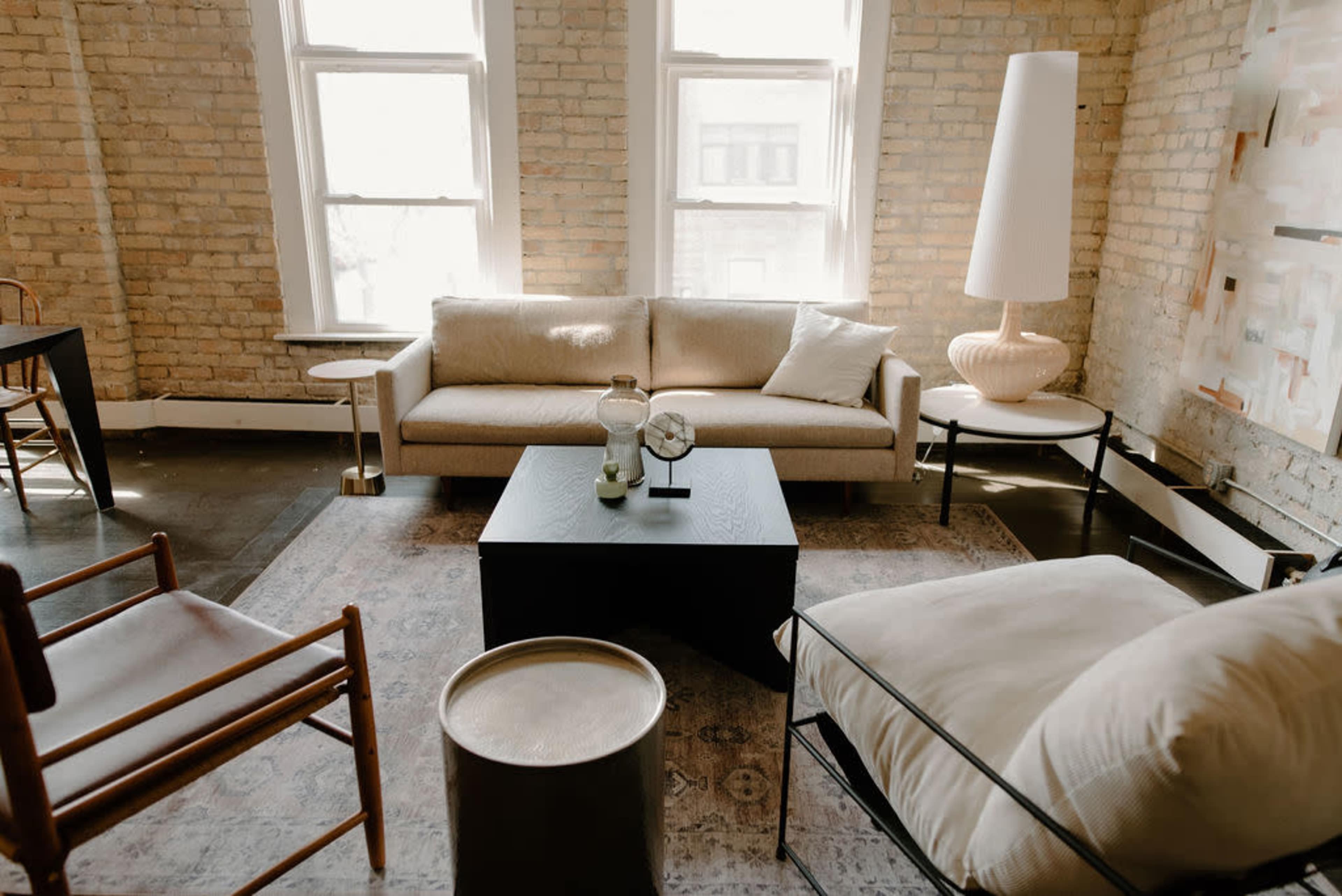 A bright living room with large windows, featuring a beige sofa, a black coffee table, a floor lamp, and a patterned area rug.