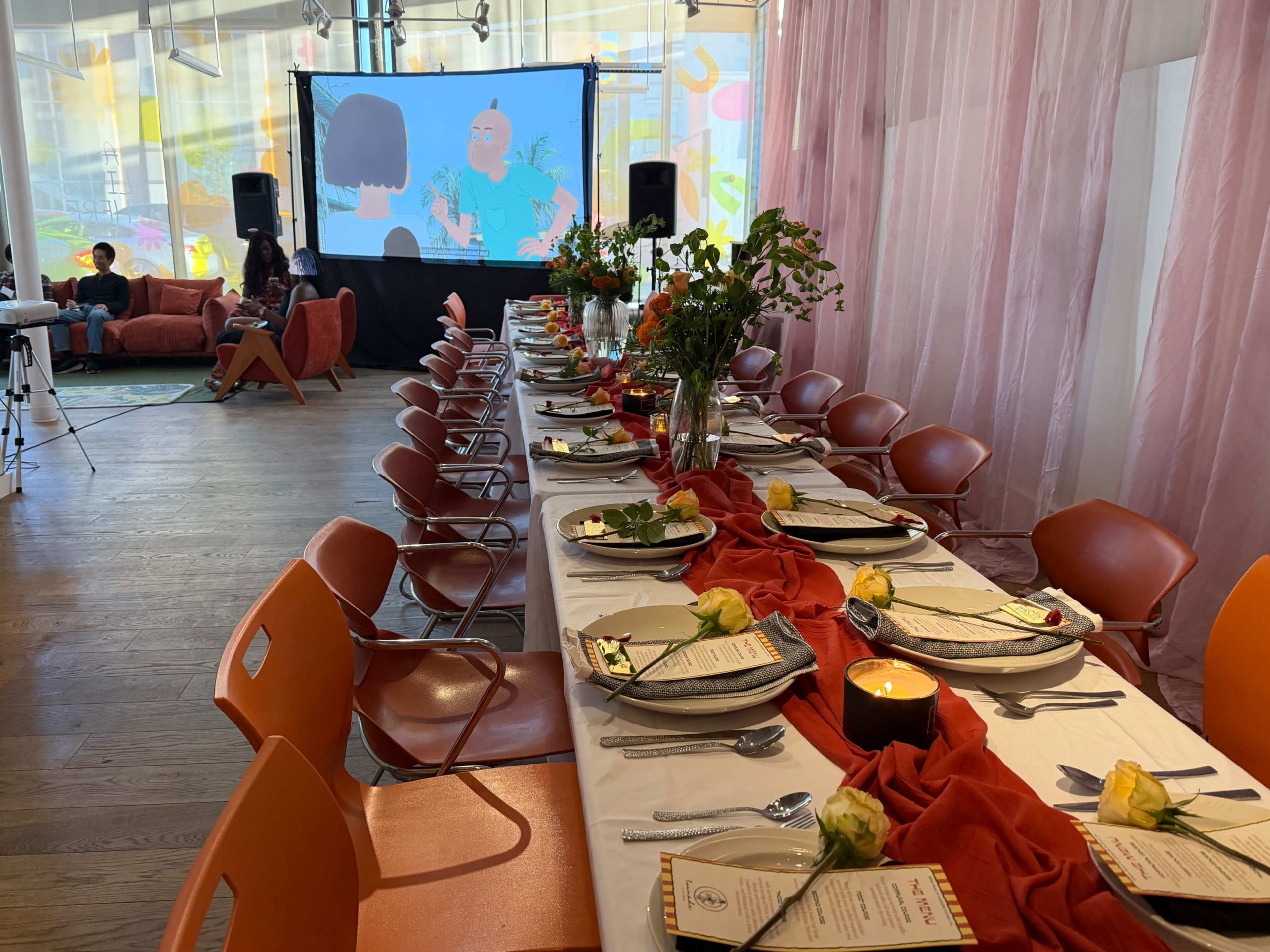 A long dining table is set with plates and cutlery, adorned with a red table runner and yellow roses, while a screen in the background displays animated characters.
