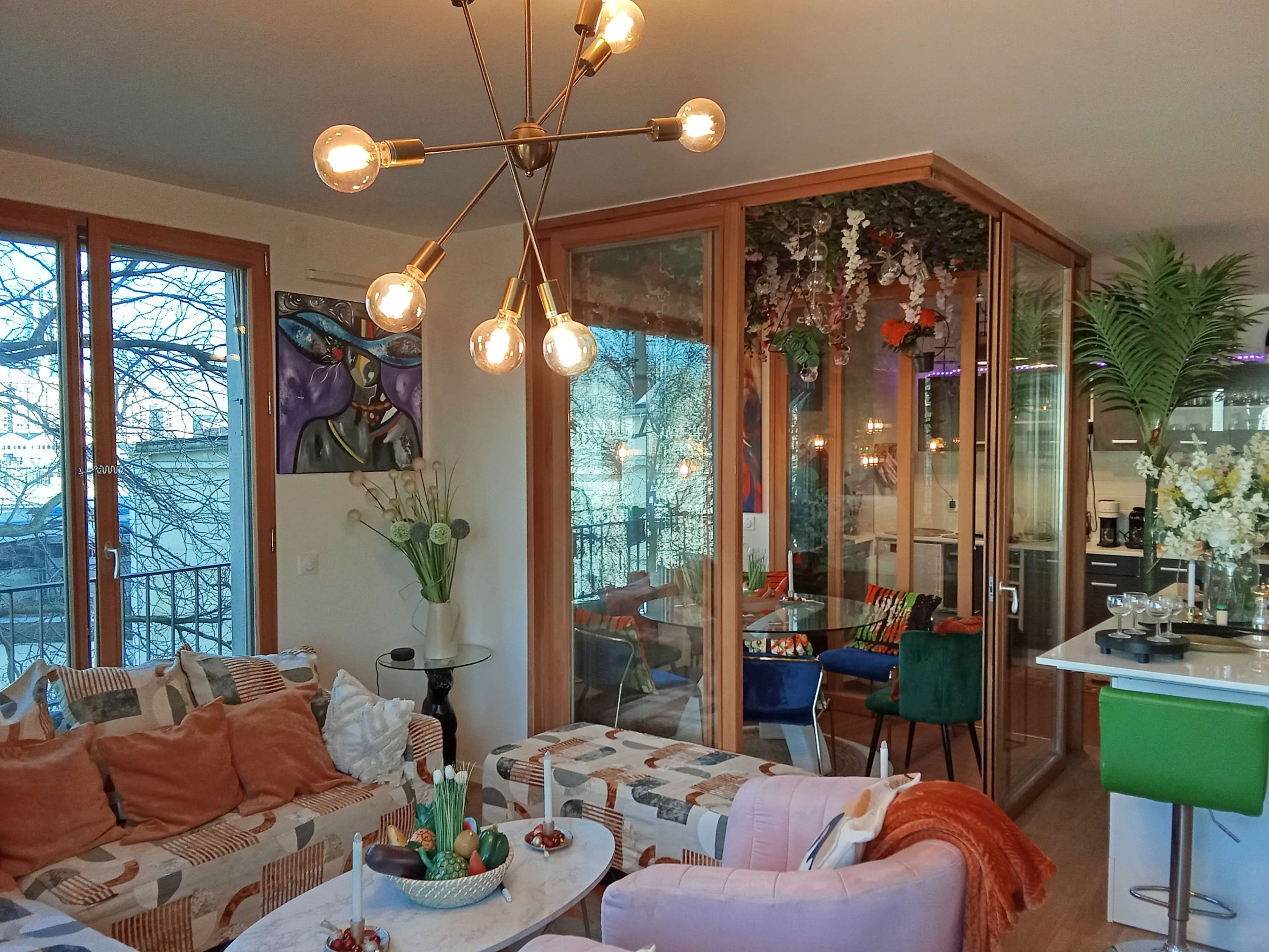 A modern living room features colorful furniture, large windows, and a dining area with plants and artwork.