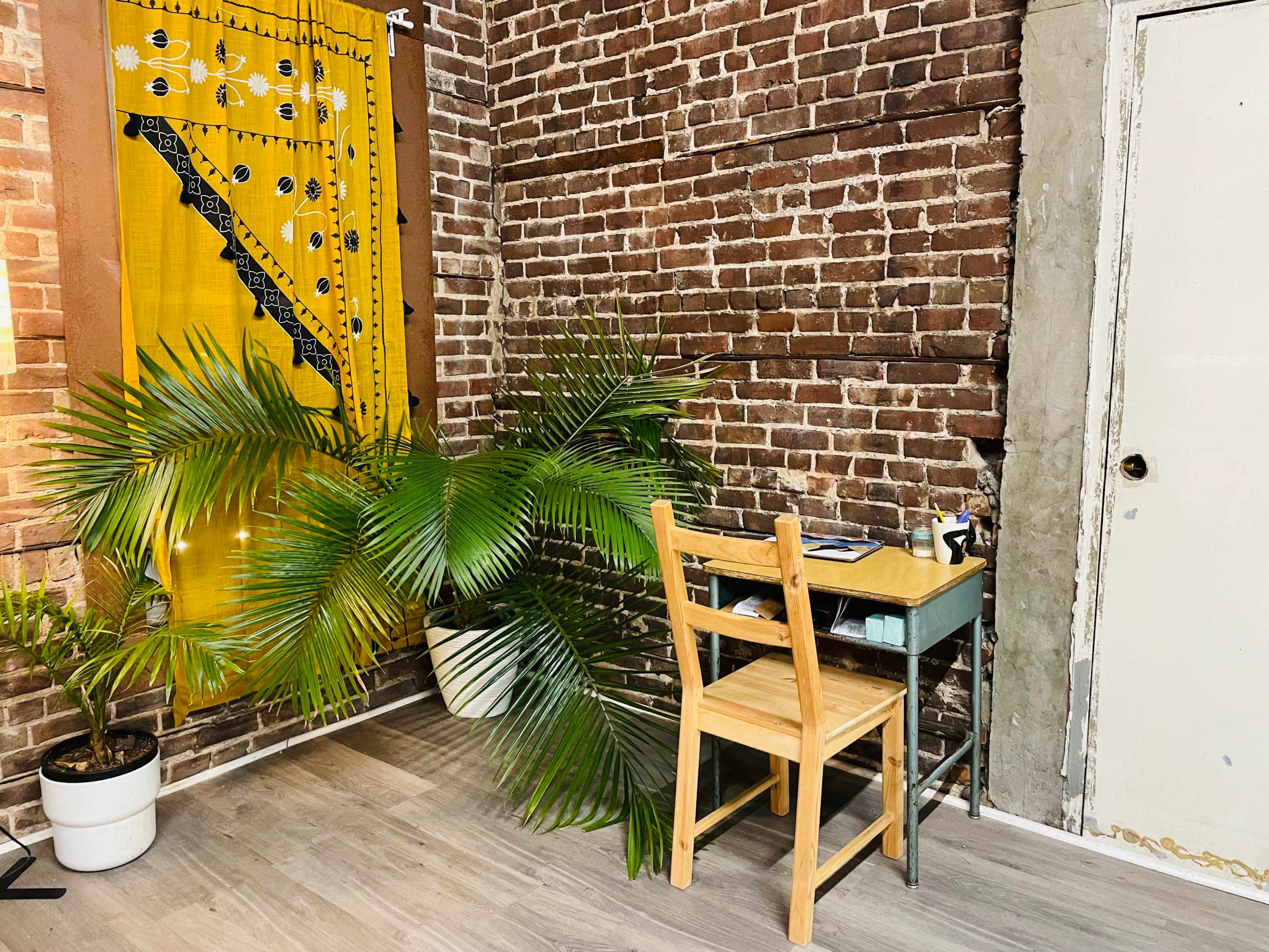 A small wooden chair sits next to a desk against a brick wall, with a large potted plant extending towards the floor.