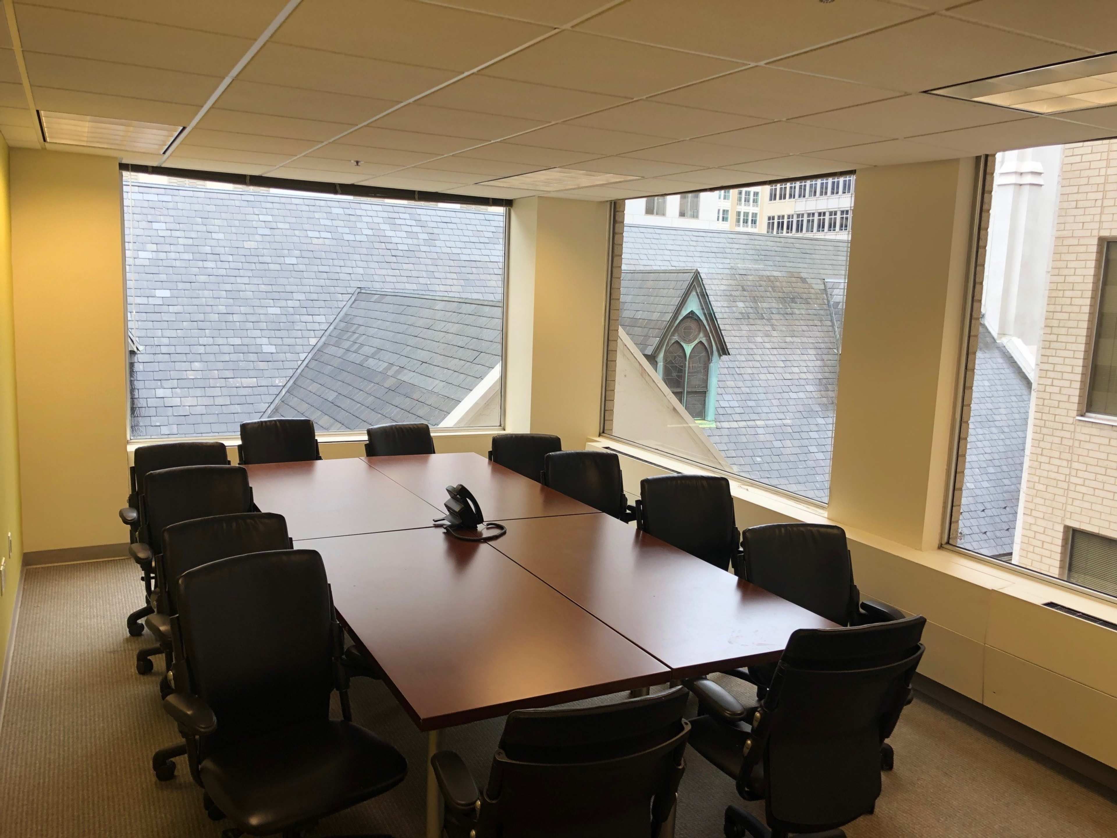 A conference room with a rectangular wooden table surrounded by black chairs and large windows overlooking rooftops.