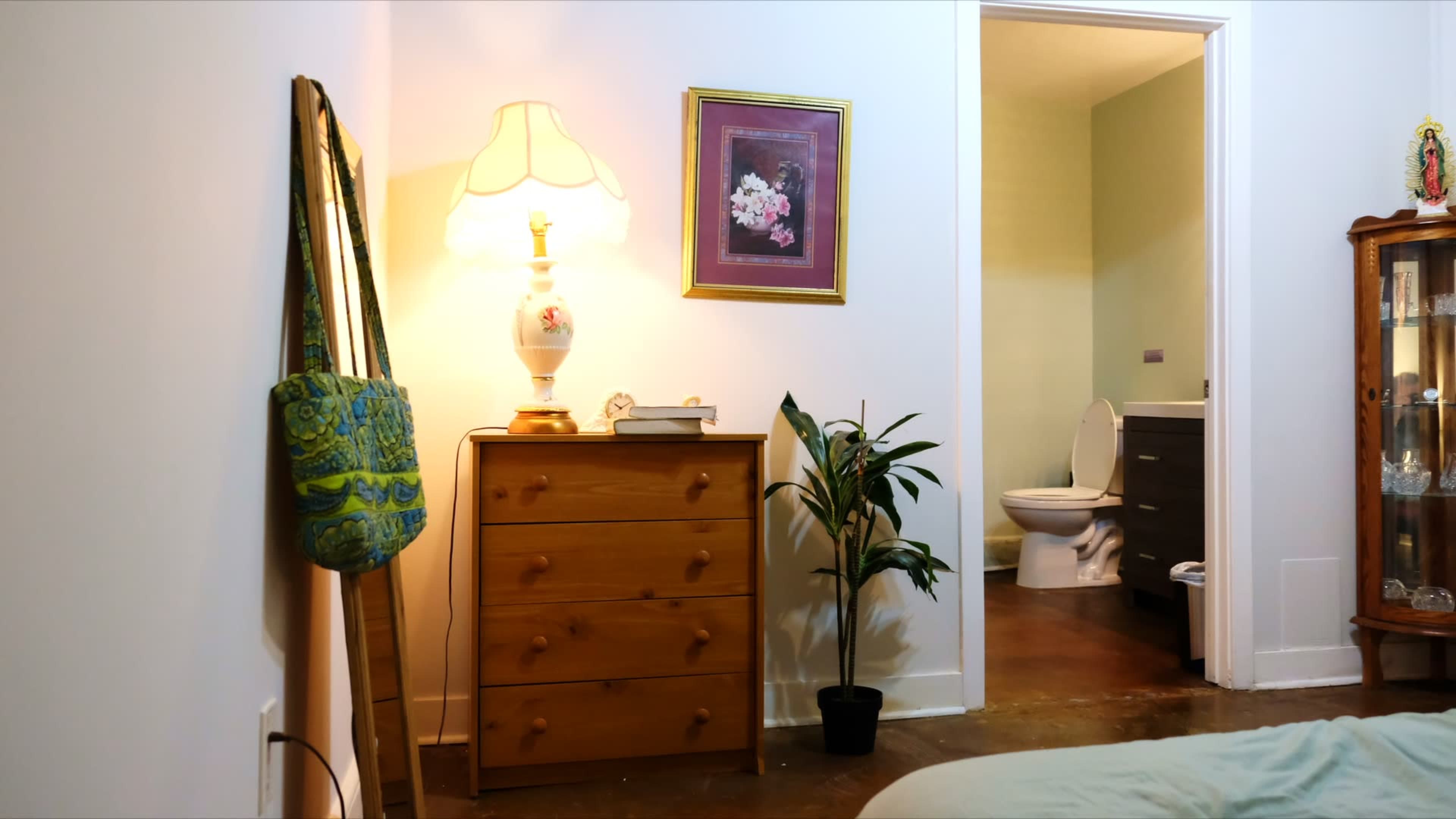 The image shows a small bedroom with a dresser, a lamp, a framed picture on the wall, and a view of a bathroom through an open door.