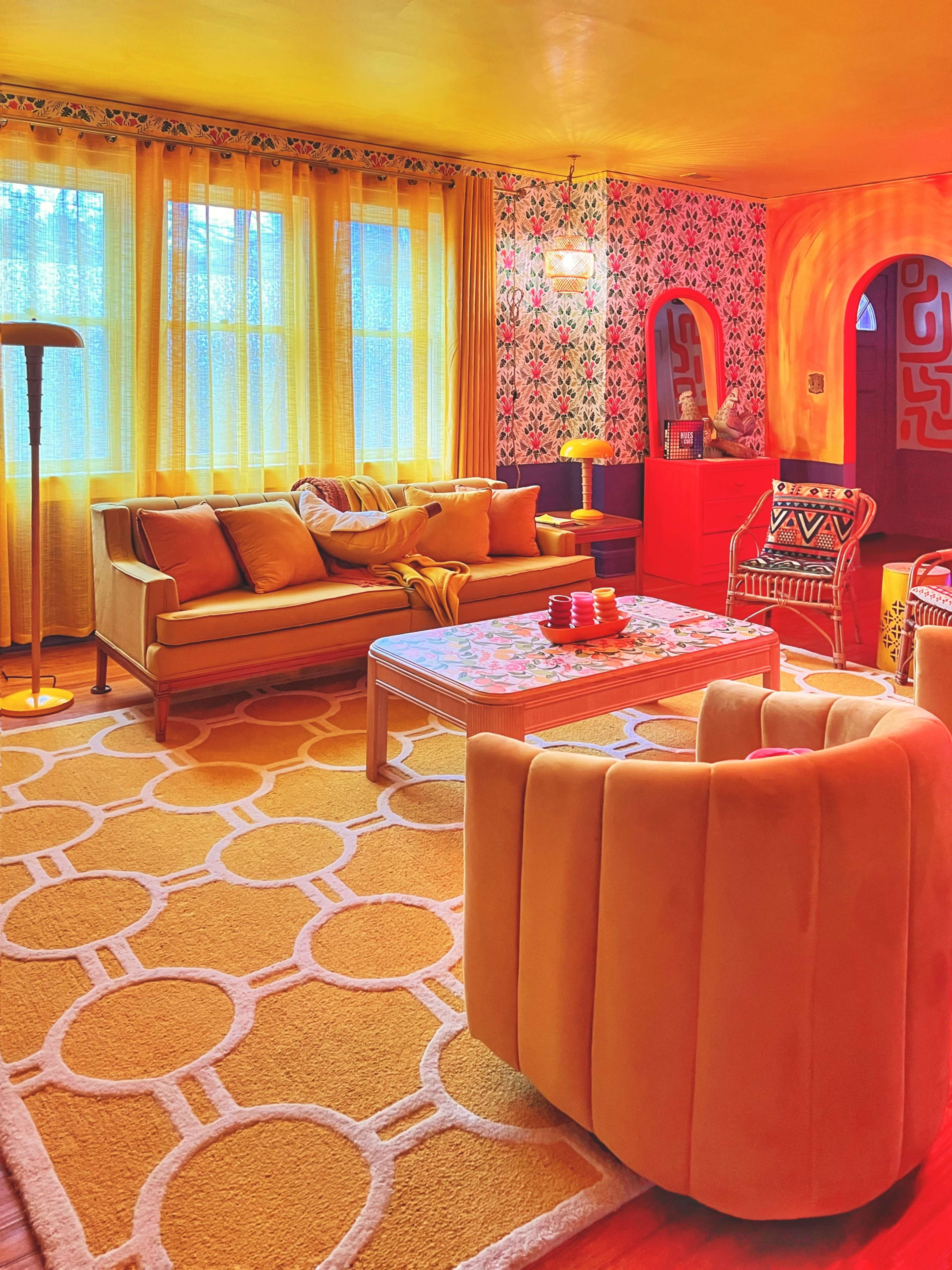 The living room features a yellow and orange color scheme with a patterned rug, a couch, armchairs, and large windows allowing natural light.
