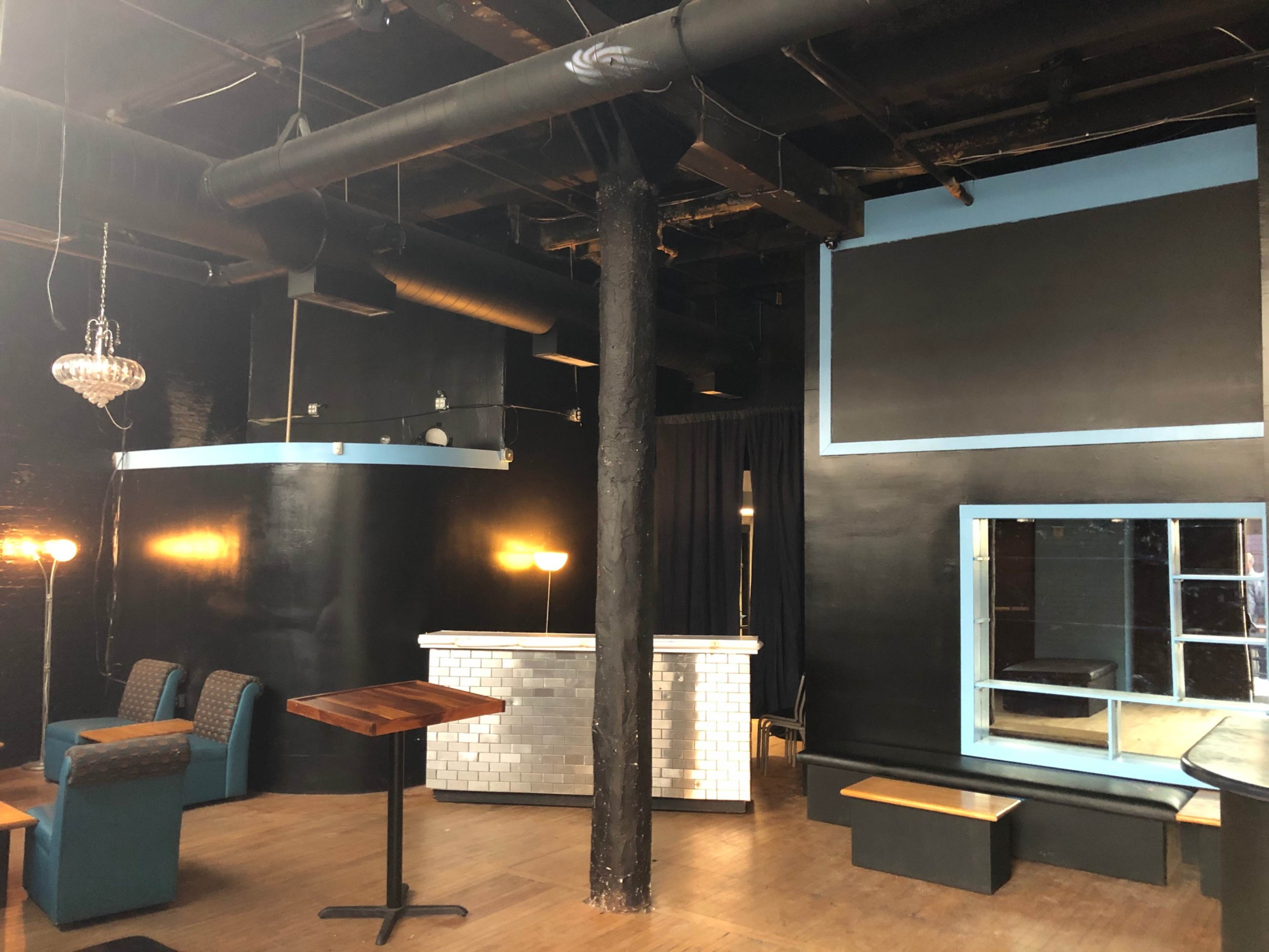 A dark room with a bar area on the left, seating arranged nearby, and a large mirror on the right wall.