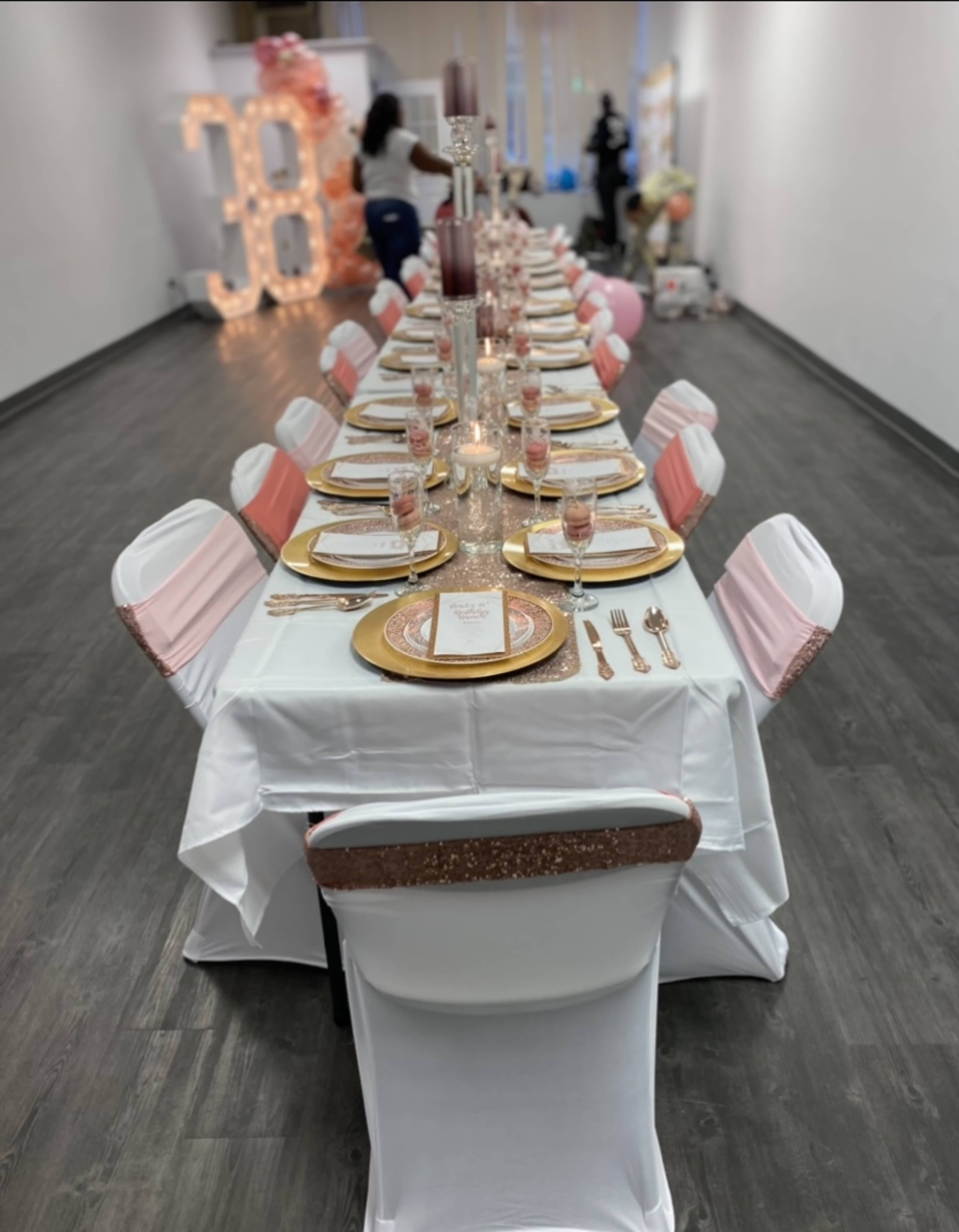 A long banquet table is set with gold and pink tableware, surrounded by chairs draped in white and pink covers, in a decorated venue.