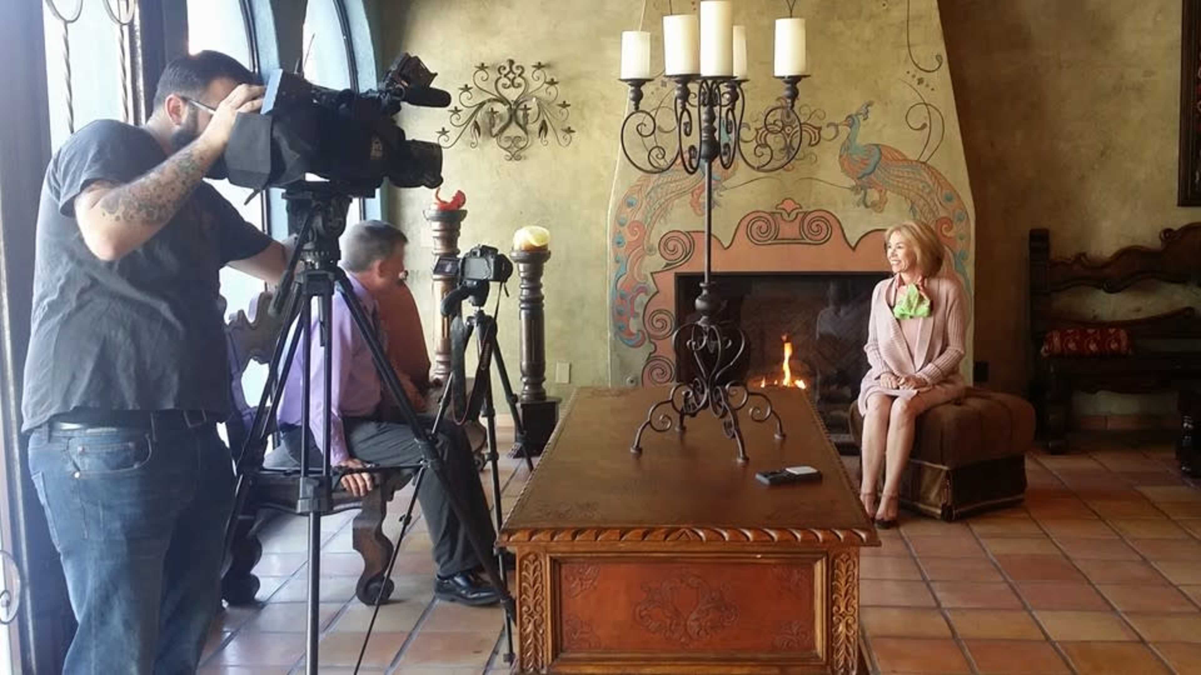 A woman sits on a couch in a decorative room while two men operate cameras nearby.