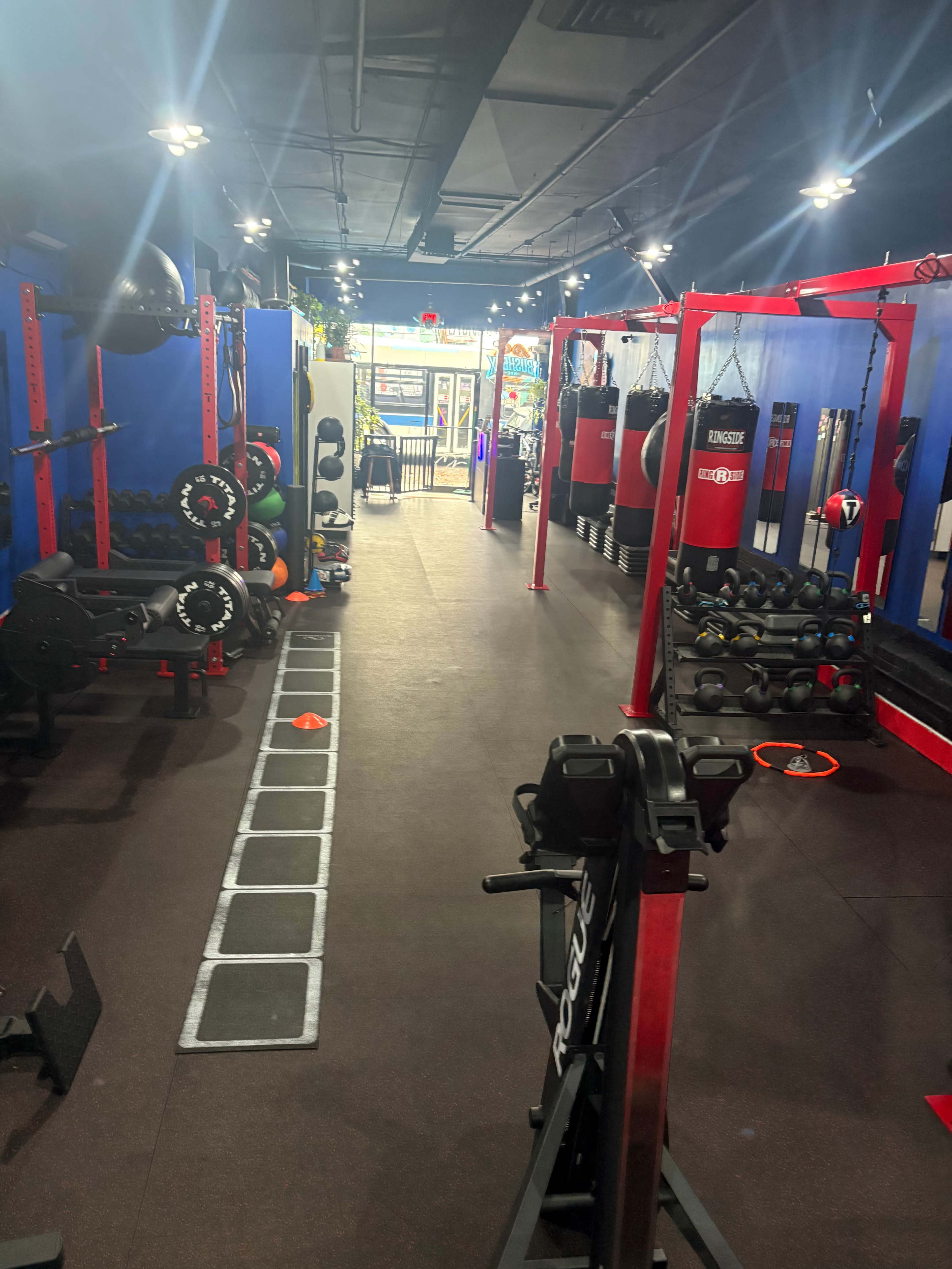 The image shows a well-equipped gym with various exercise machines, weights, and fitness accessories arranged along a long, narrow space.