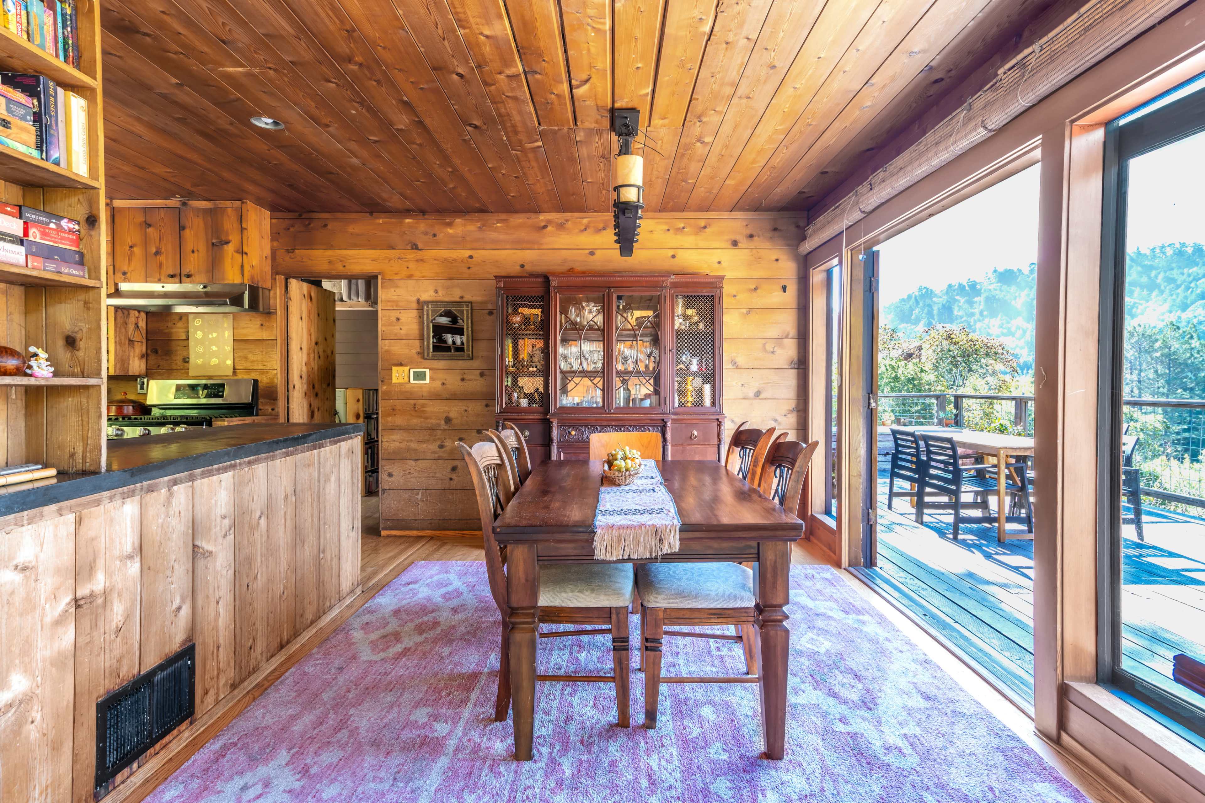 The interior of a wooden dining room features a long table set for meals, a glass-fronted cabinet displaying dishware, and large windows that open to an outdoor deck with a view of greenery.