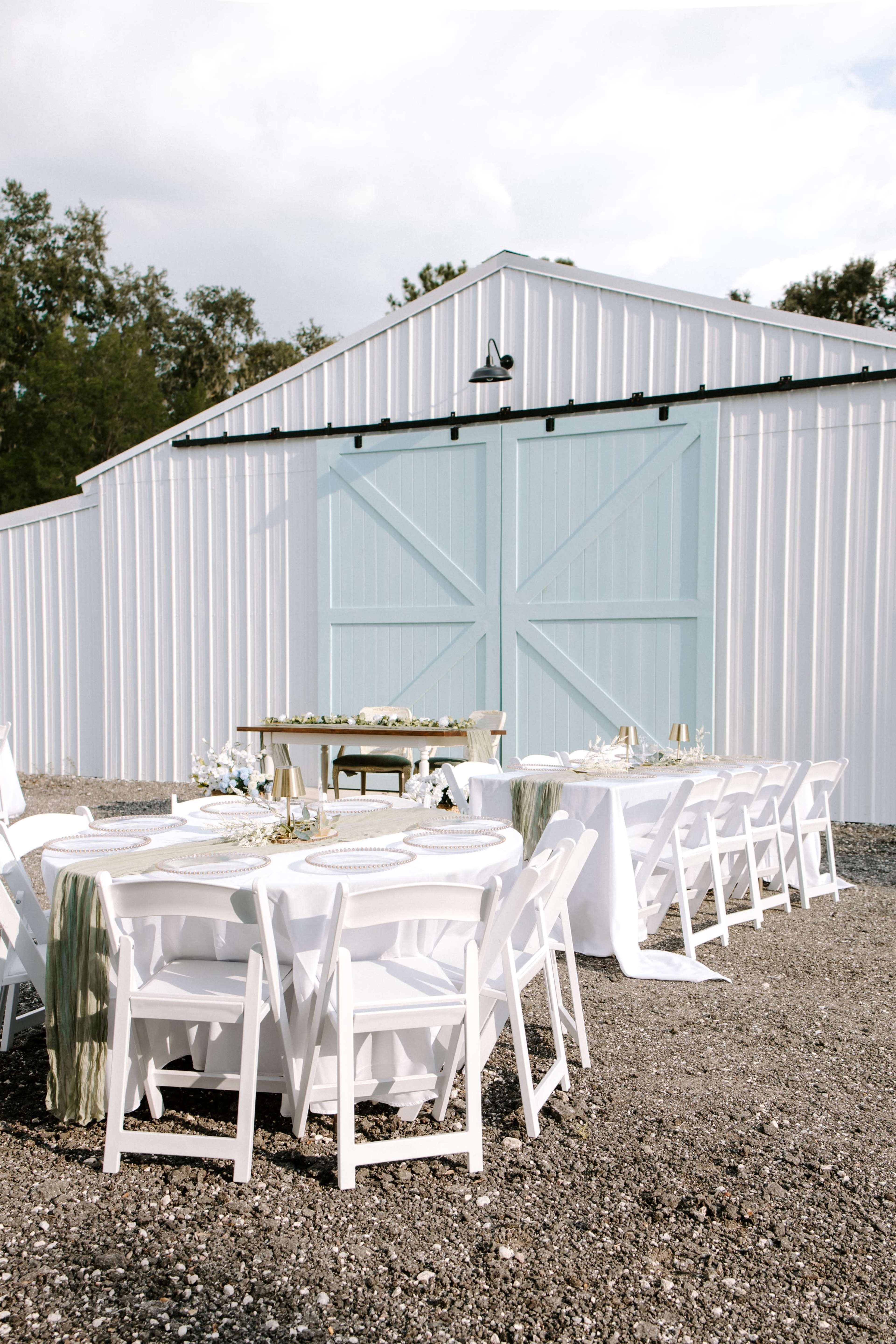 A set of white tables and chairs are arranged outside in front of a large blue barn door, with a small table decorated with flowers nearby.