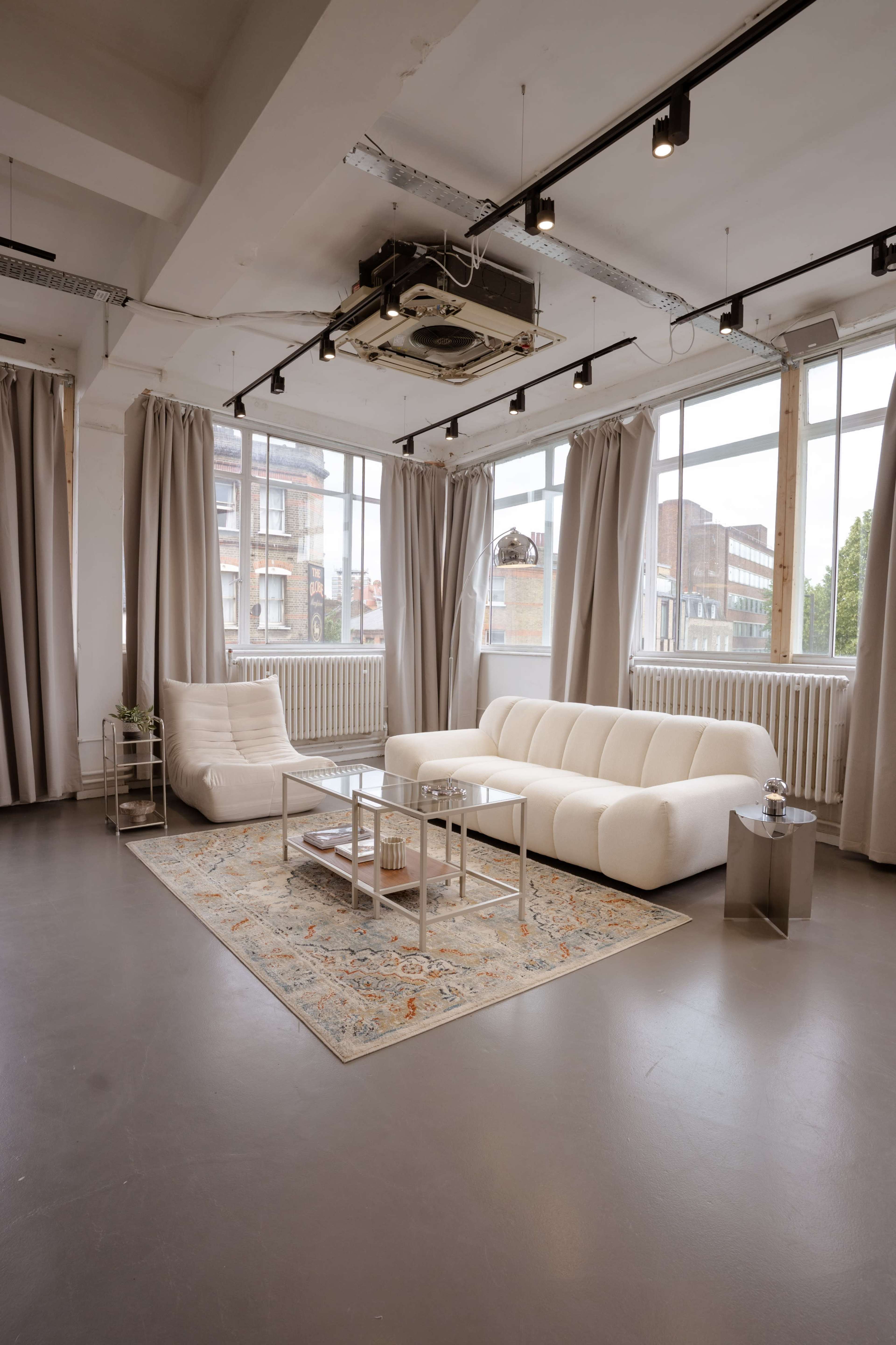 The image shows a bright and spacious interior with large windows, featuring a white sofa, a glass coffee table, and a patterned area rug on the floor.