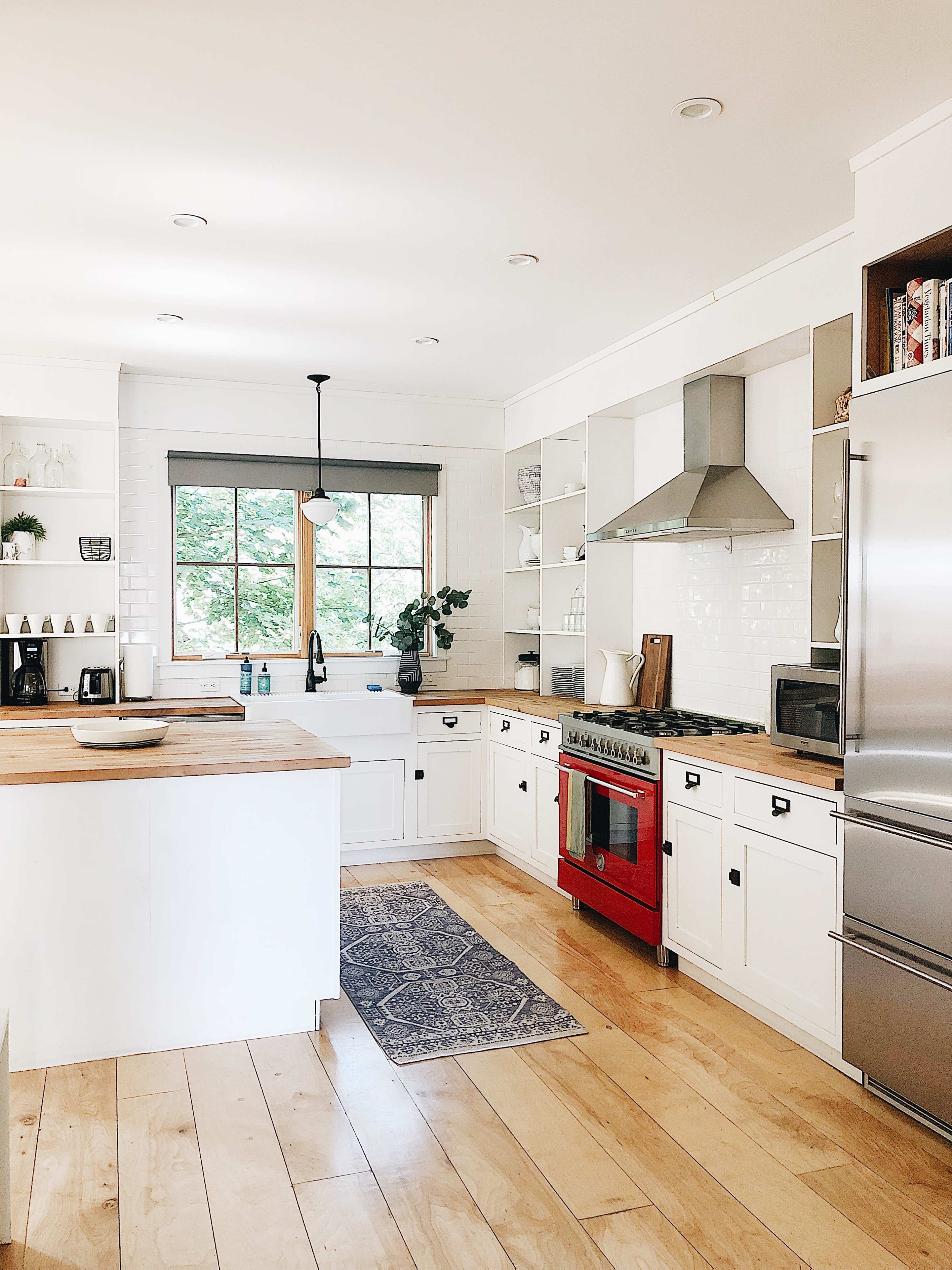 A bright kitchen features white cabinetry, a wooden island, a red stove, and large windows allowing natural light to illuminate the space.
