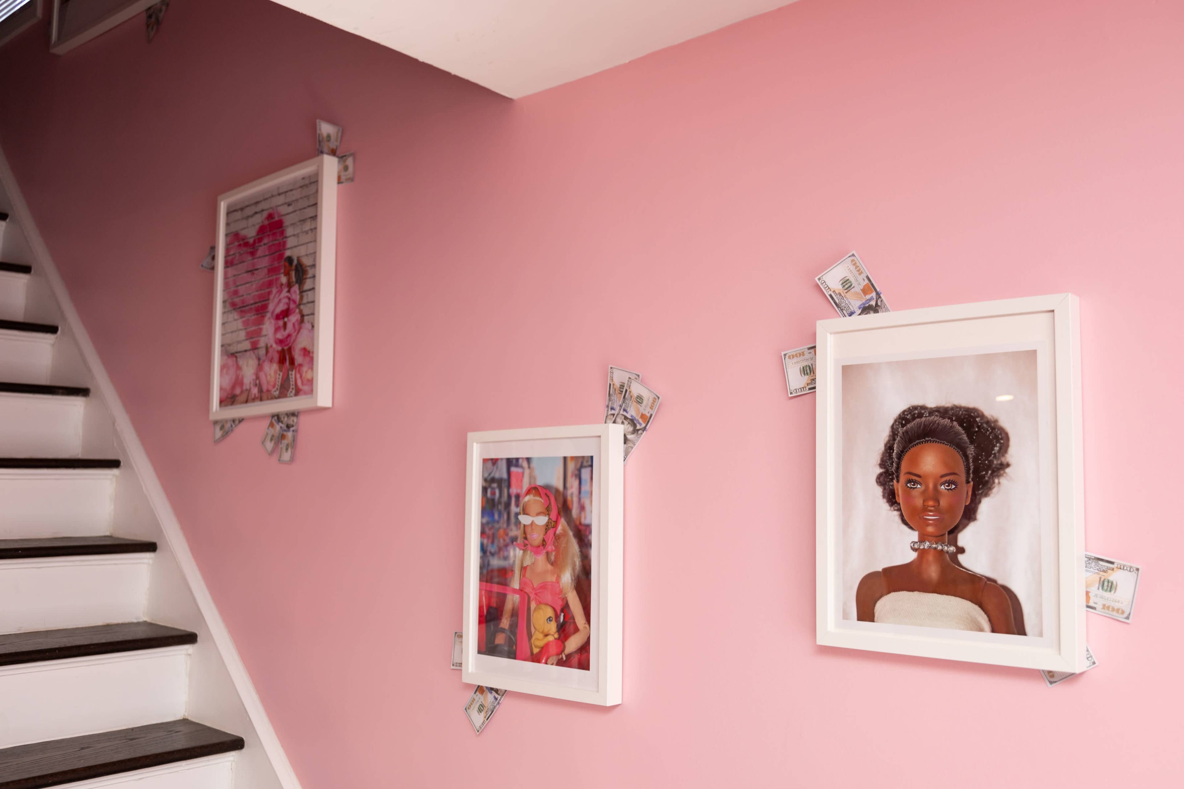 Three framed images of dolls are mounted on a pink wall, with dollar bills arranged around the edges of each frame.