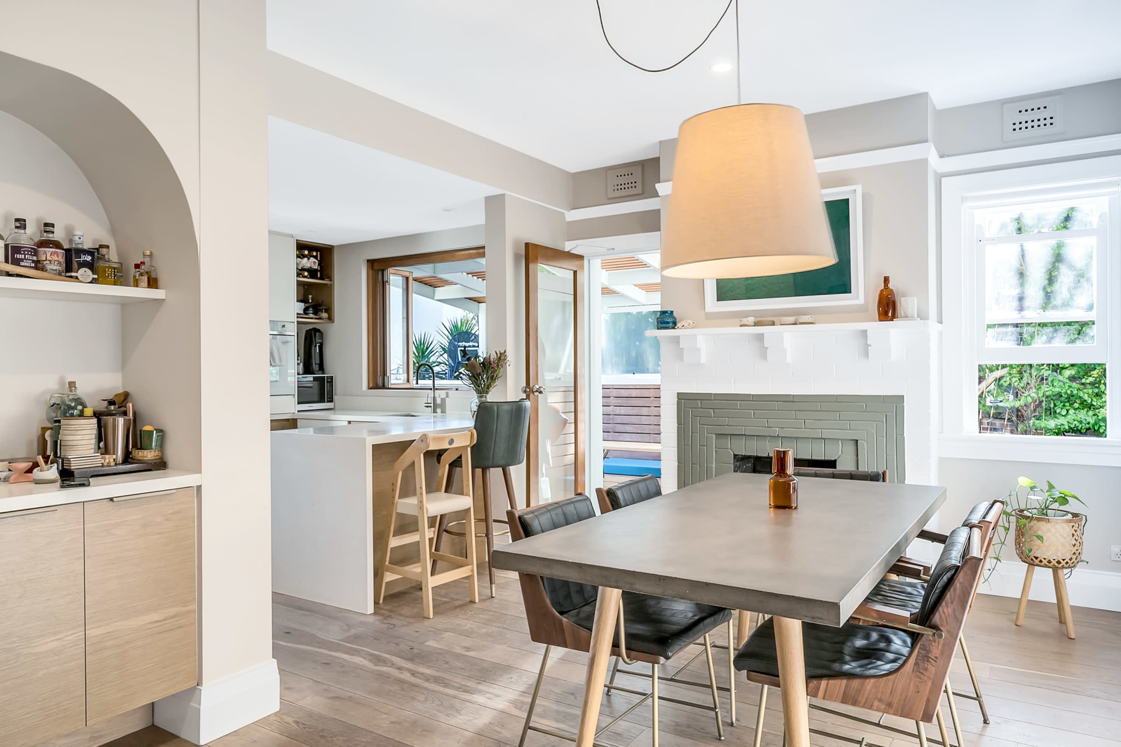 A modern kitchen and dining area with a concrete dining table, wooden chairs, and a green fireplace, featuring large windows that provide natural light.