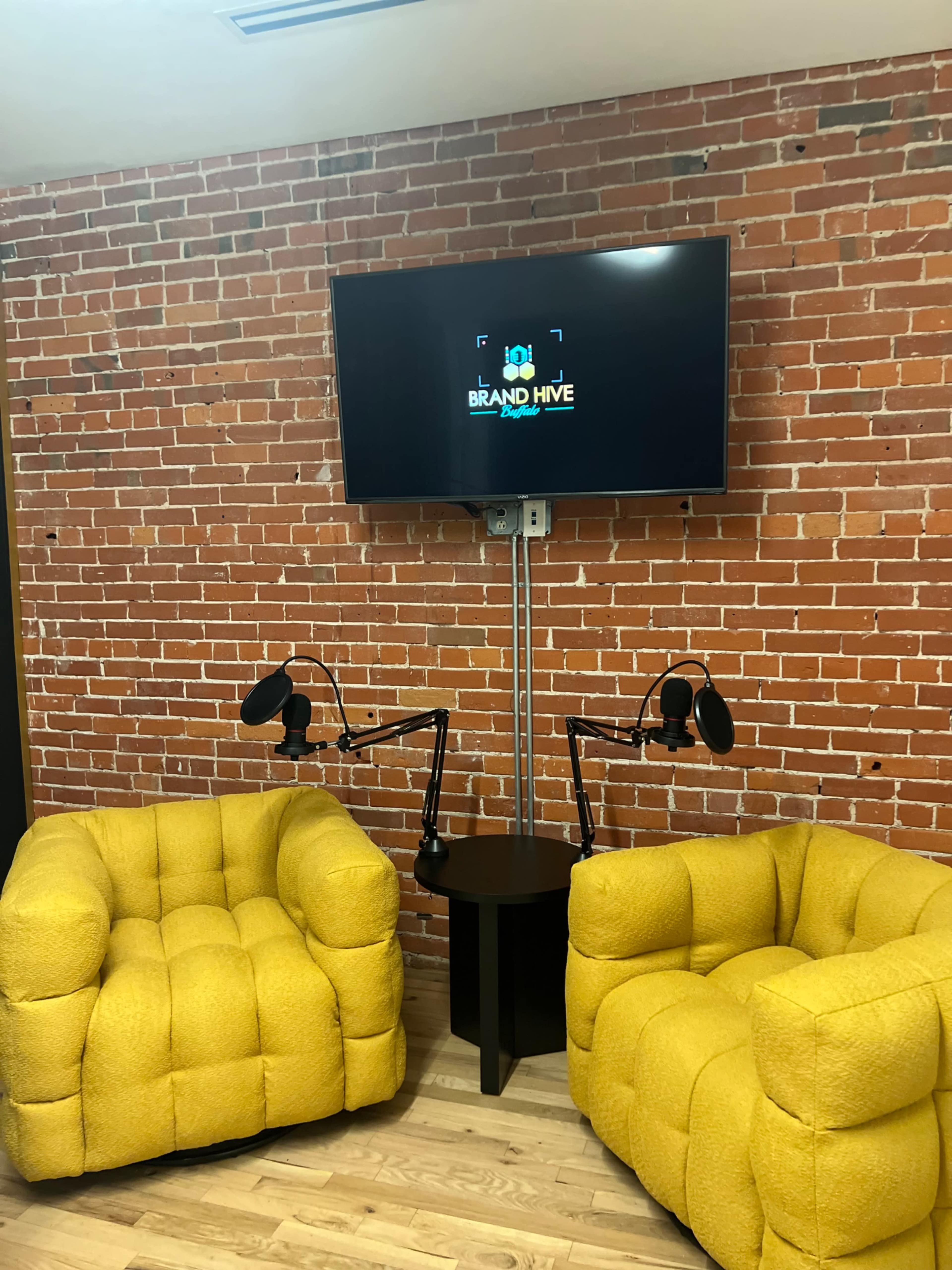 Two yellow upholstered chairs positioned beside a small black table, with a television mounted on a brick wall displaying the logo for "Brand Hive."