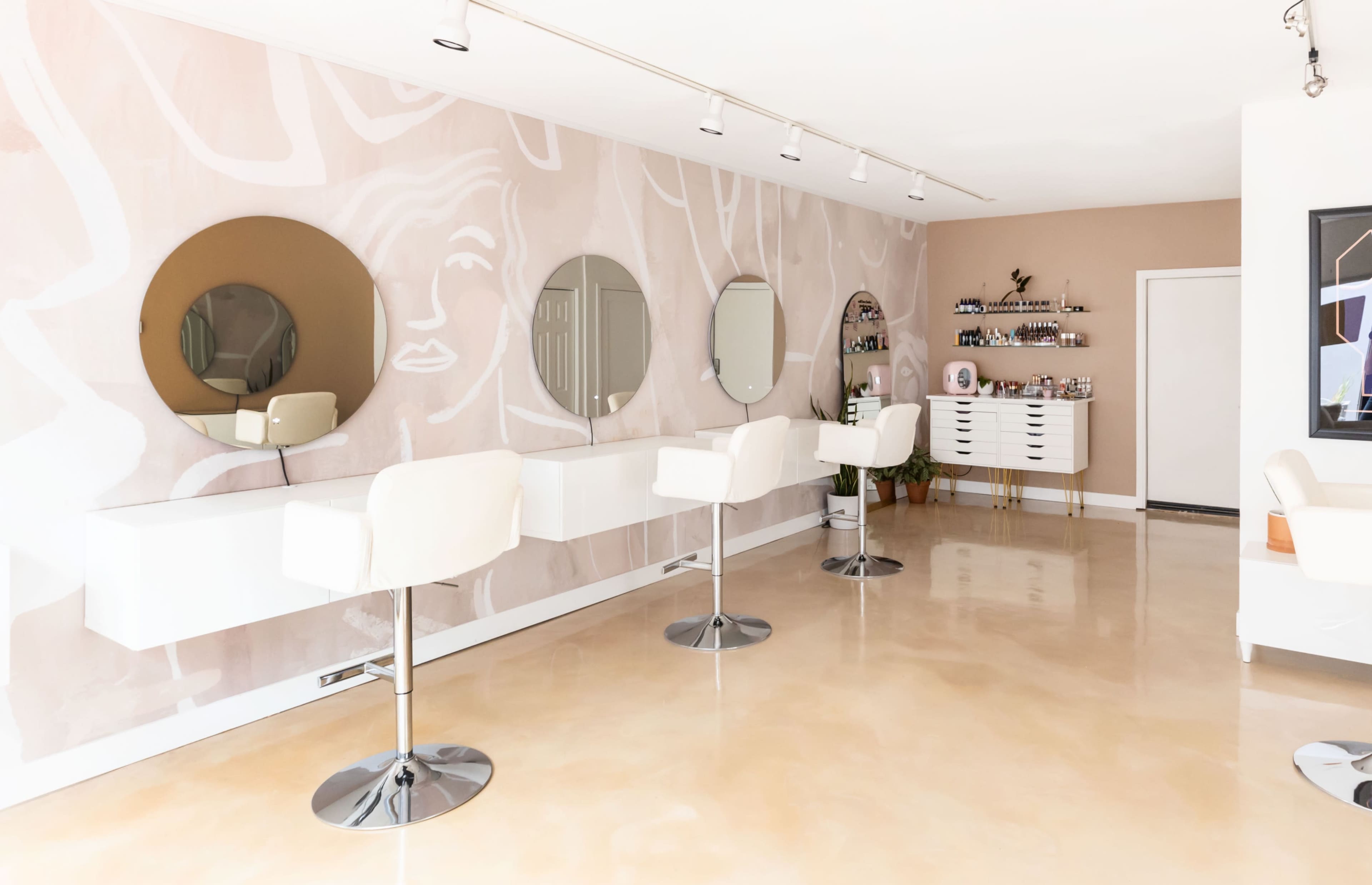 A modern salon interior with three white bar stools and large round mirrors along one wall, while a minimalist shelving unit holds various beauty products on the opposite side.