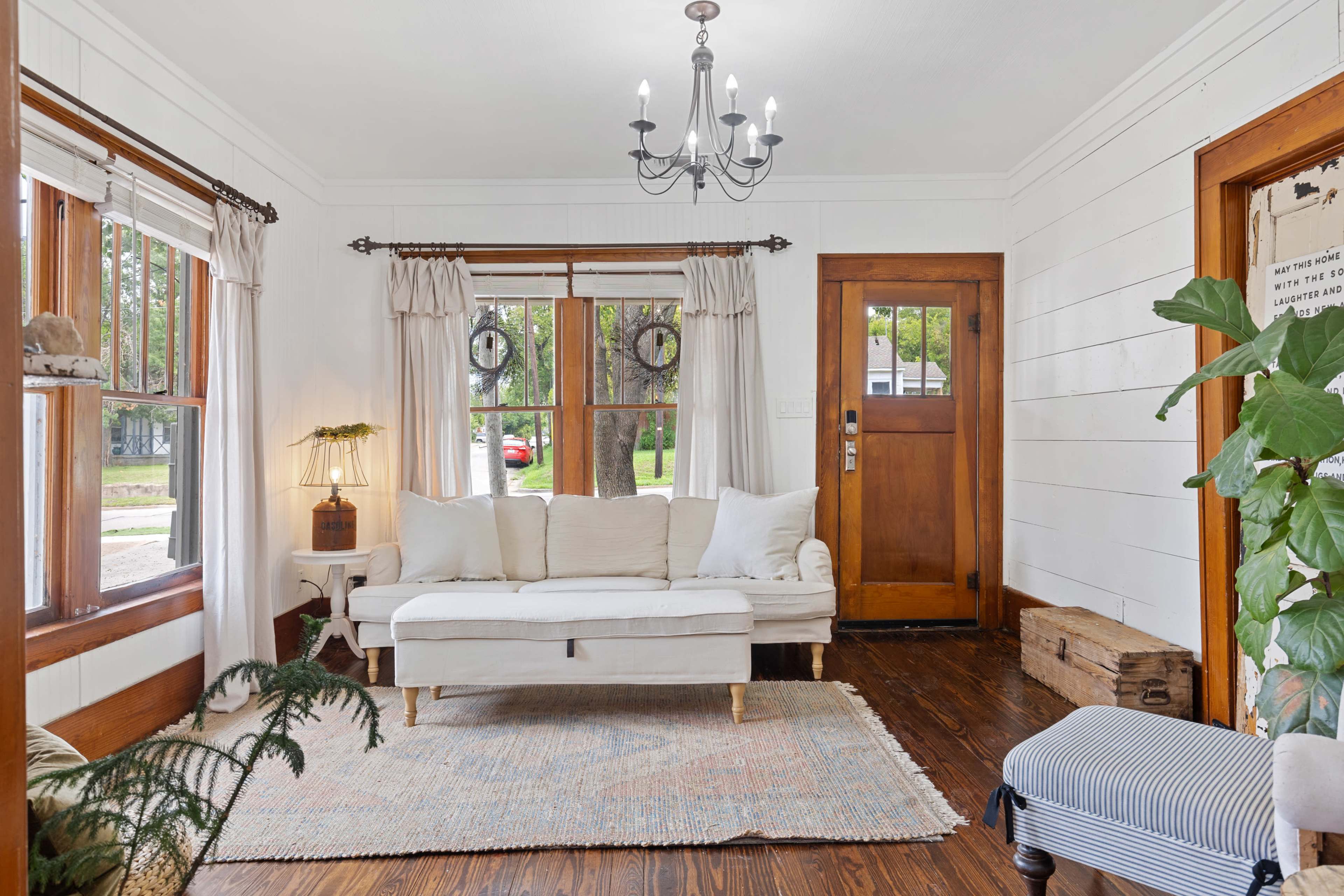 The living room features a white sofa, a wooden door, and a chandelier, along with large windows framed by curtains.