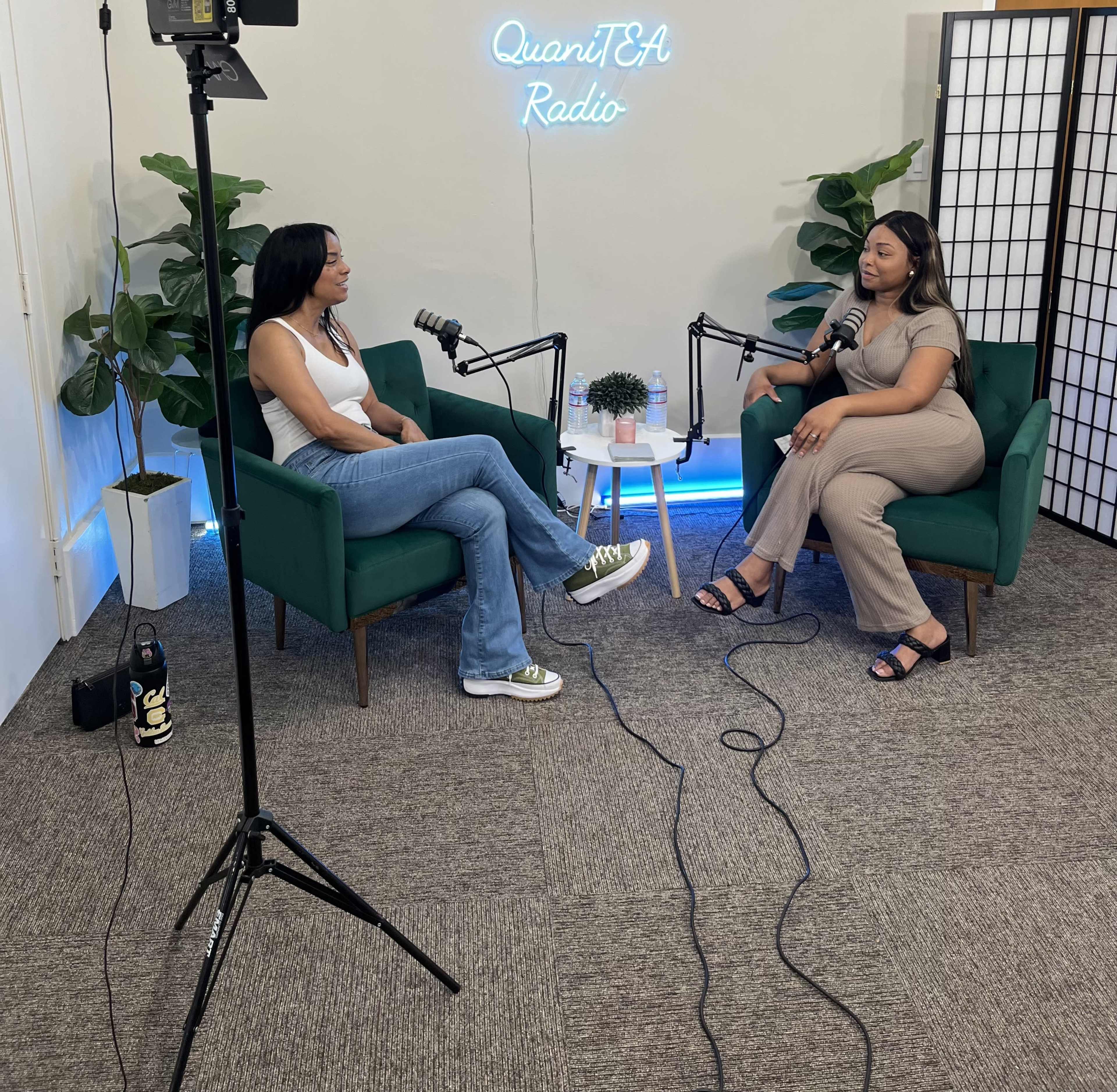 Two women are sitting in green chairs across from each other in a podcast studio, with microphones and plants in the background.