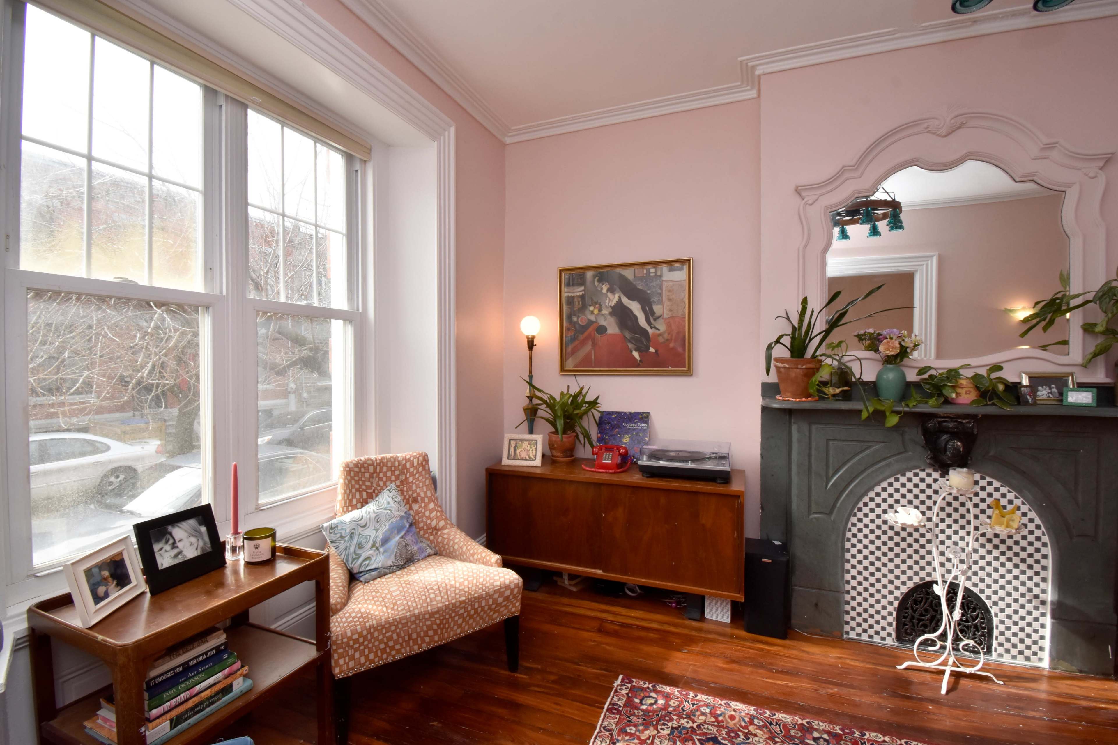 A cozy corner of a living room features a patterned chair next to a wooden table, framed by large windows and a decorative fireplace with plants and artwork.