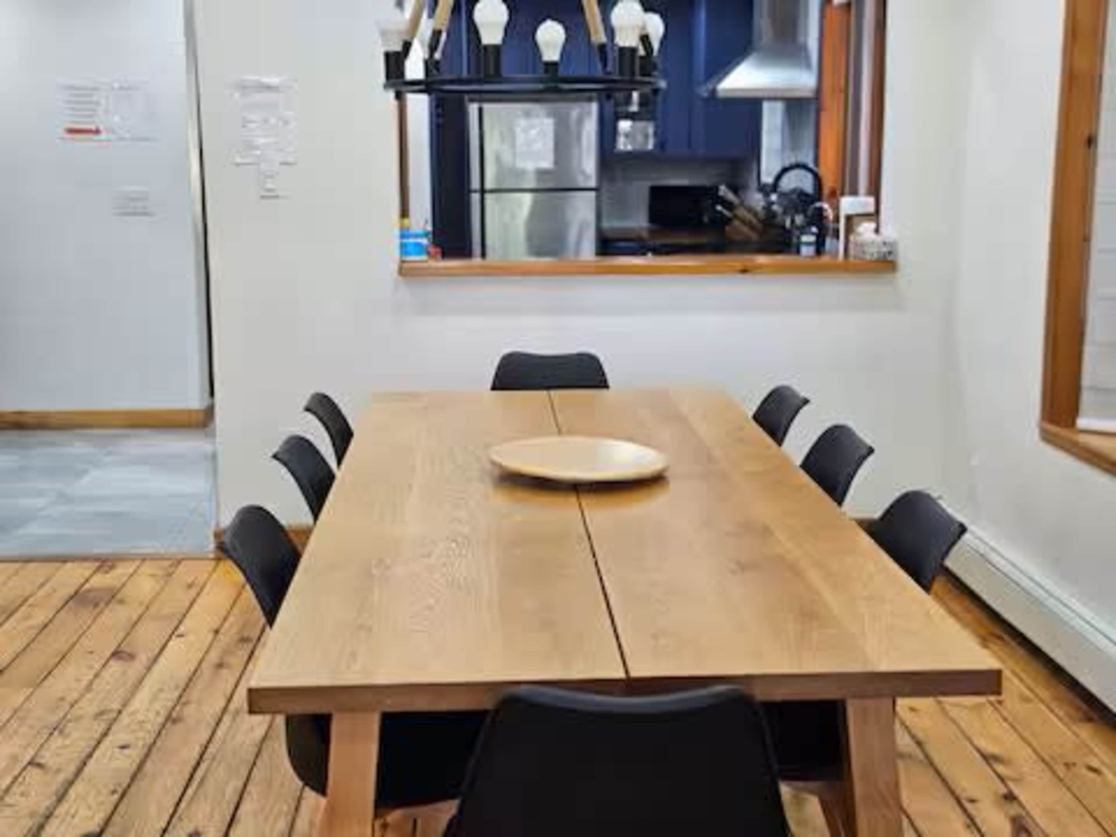 A wooden dining table with a round centerpiece is surrounded by black chairs in a well-lit dining area that connects to a modern kitchen.
