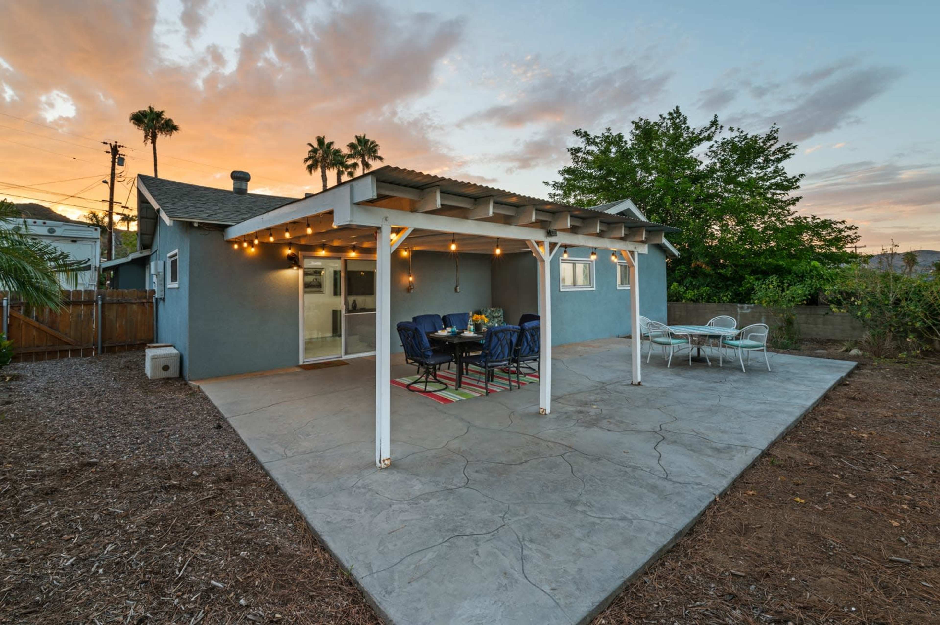 A patio area with a dining table and chairs is set under a wooden structure, surrounded by a garden and trees at sunset.