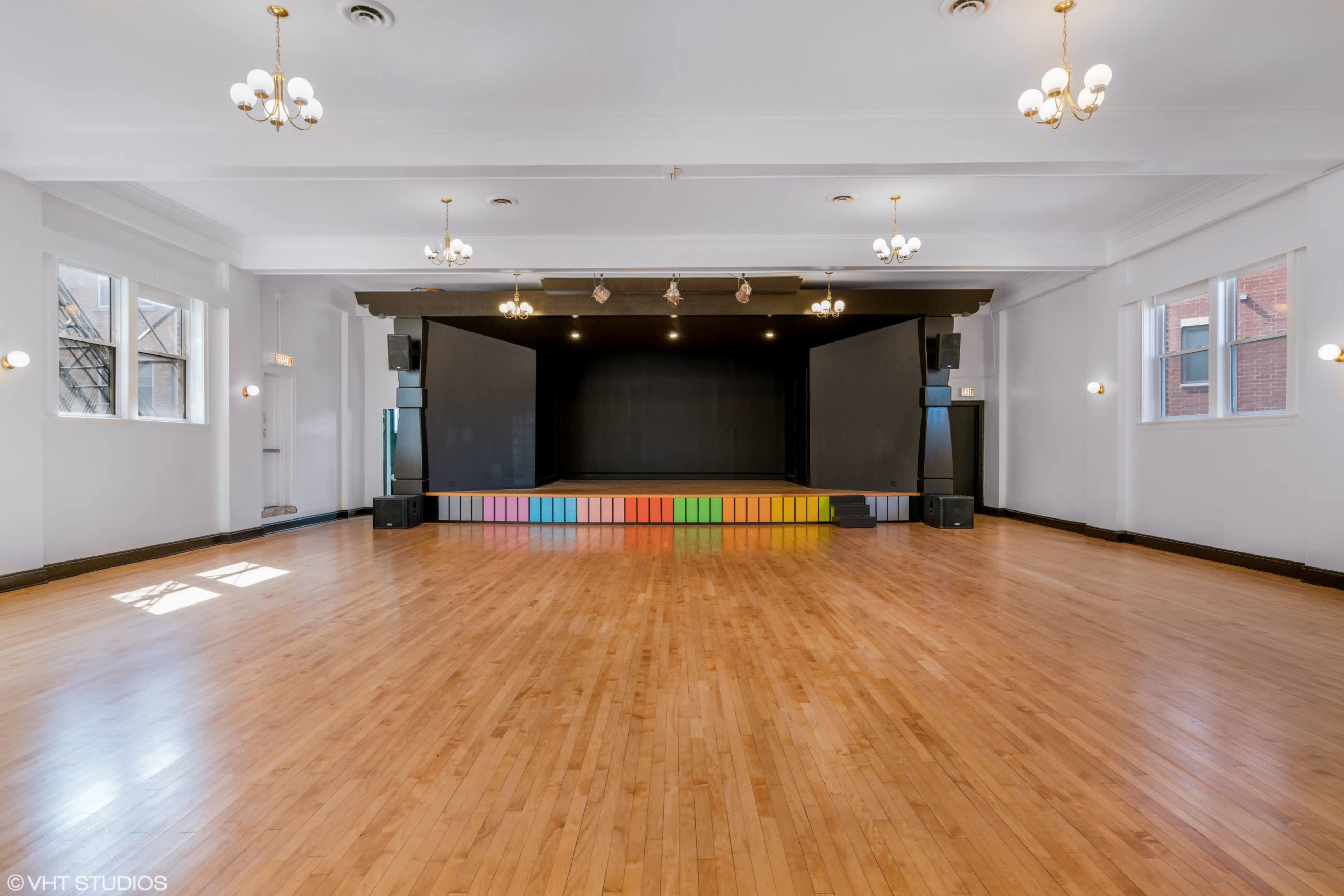 A spacious, empty hall with polished wooden floors, a stage at the front featuring colorful steps, and bright lighting from overhead chandeliers.