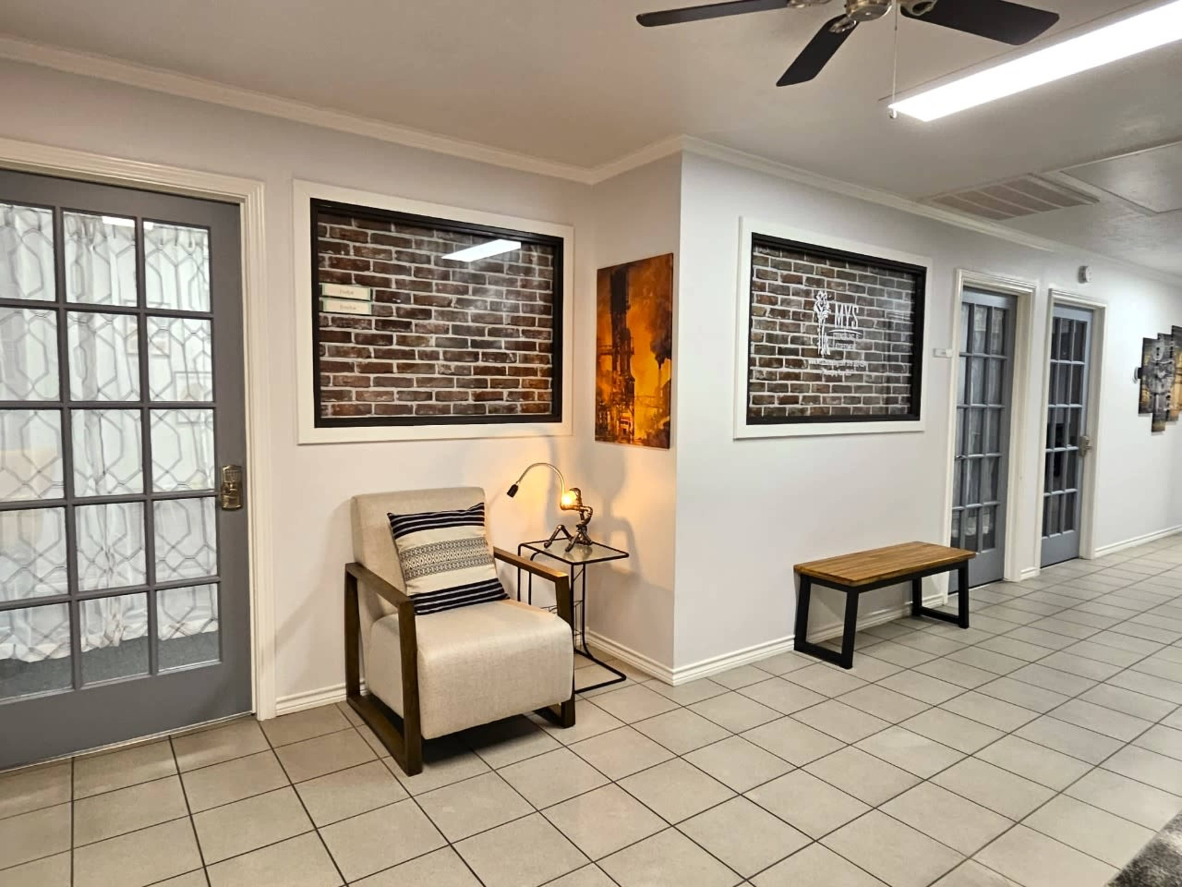 The image shows a hallway with tiled flooring, featuring a chair and side table next to a wall adorned with brick and artwork, flanked by two sets of doors.