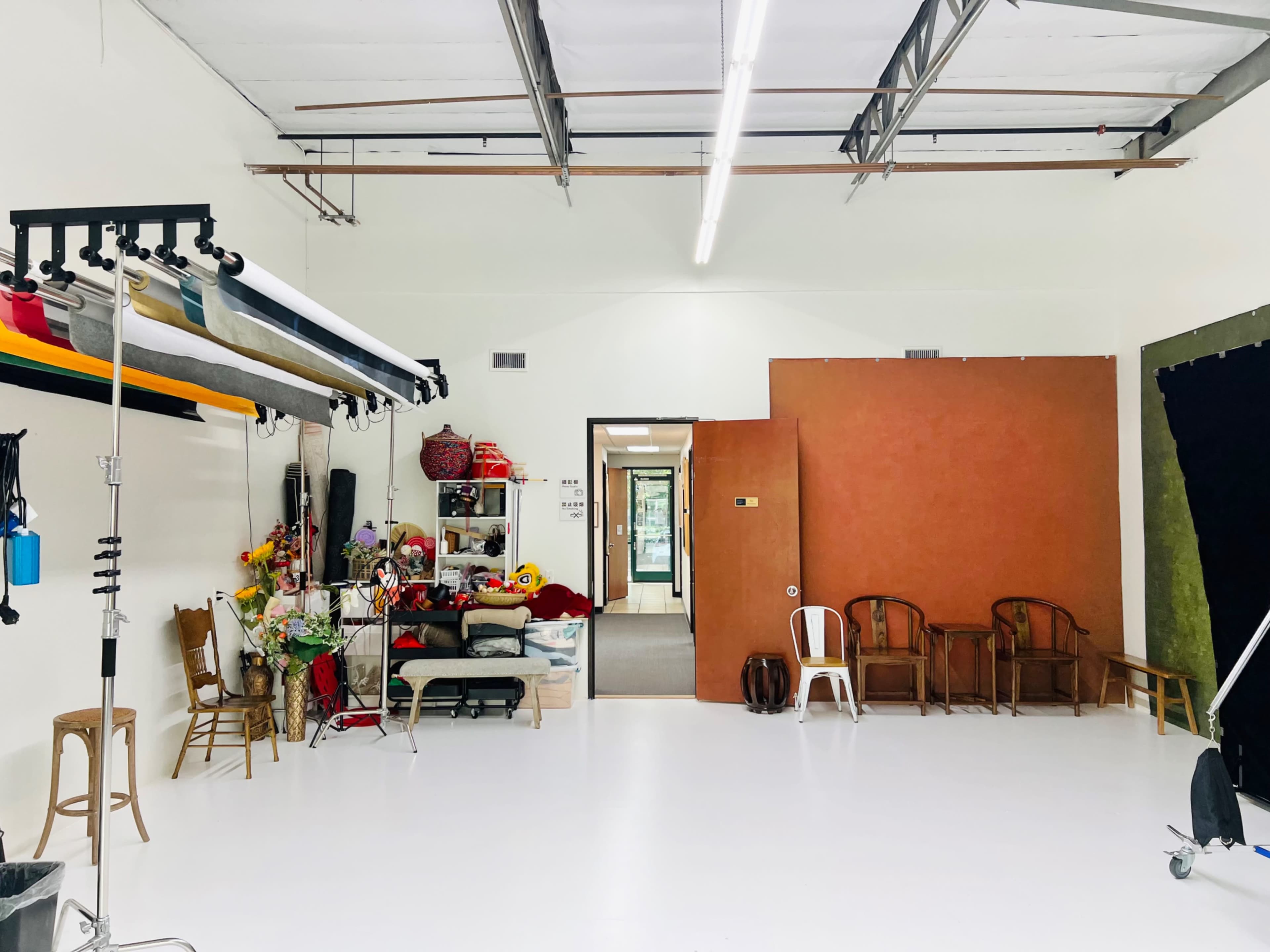 The image shows a spacious studio with a clean white floor, various props and furniture along one side, and a large brown wall at the back.