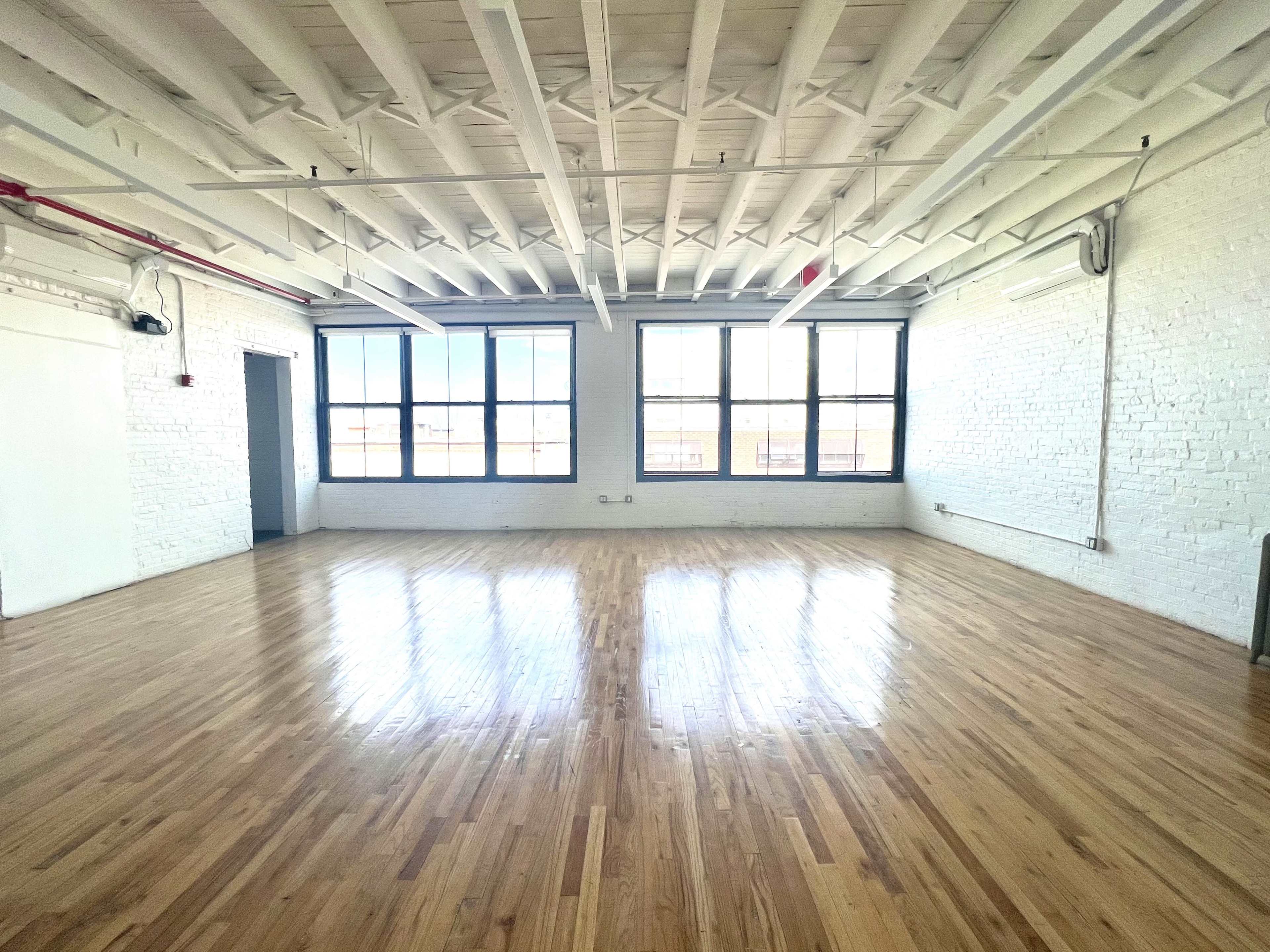 The image shows a spacious, empty room with hardwood floors and large windows along one wall, illuminated by natural light.