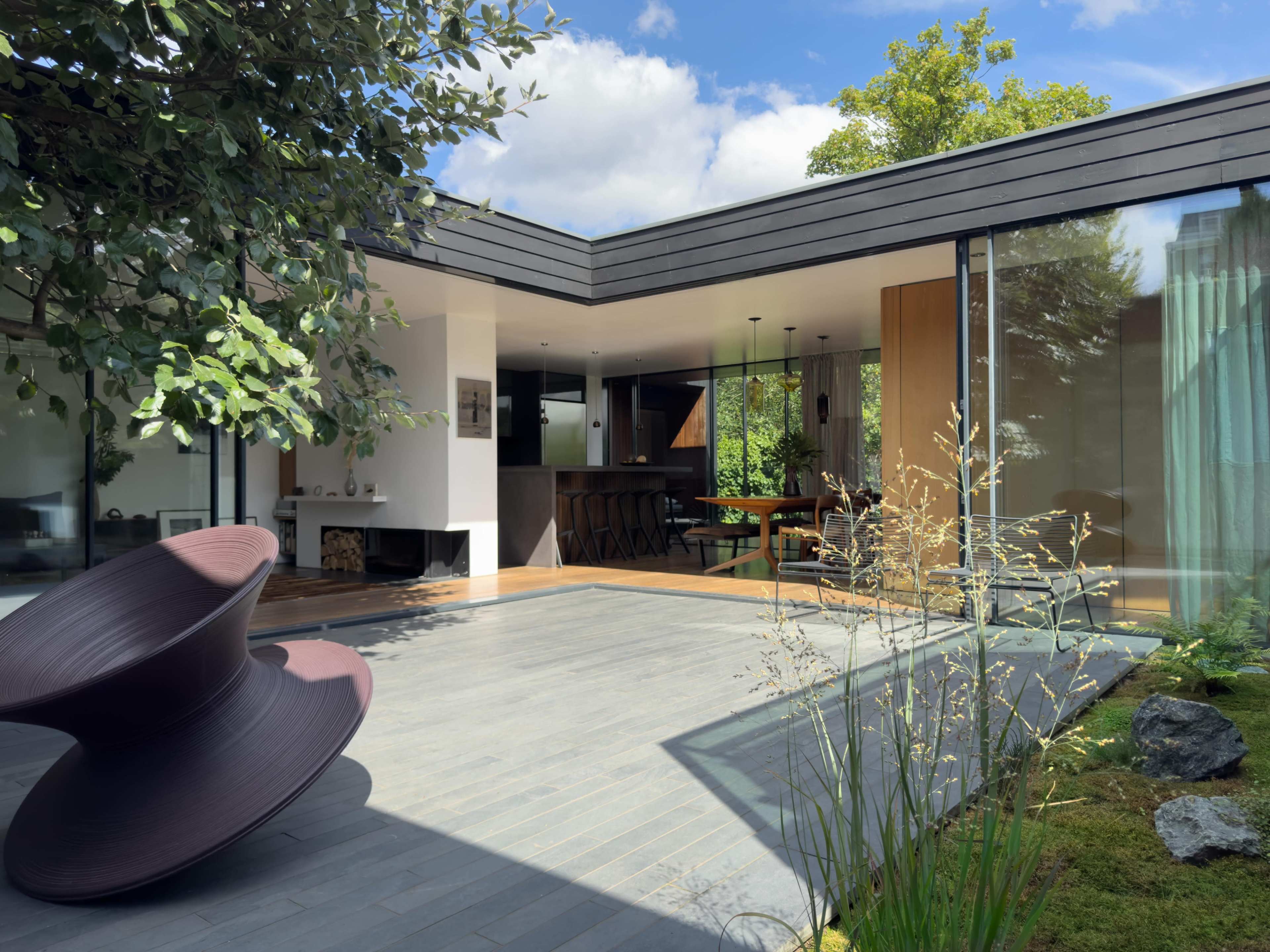 The image shows a modern home with a central courtyard featuring a sculptural seating piece, surrounded by glass walls and greenery.